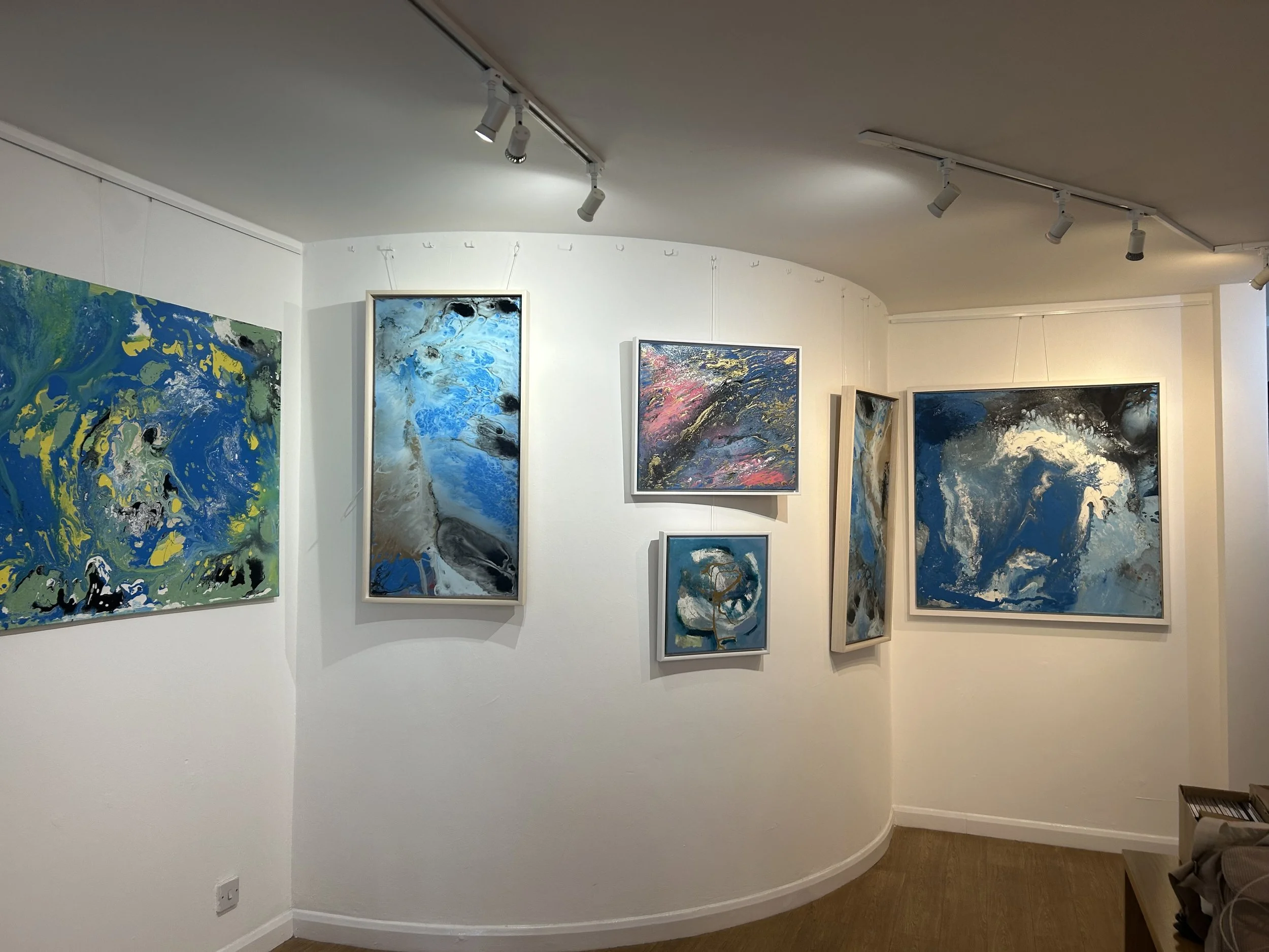 Art gallery wall featuring abstract paintings in shades of blue, black, white, and pink, displayed in white frames and on the gallery's white walls with track lighting.