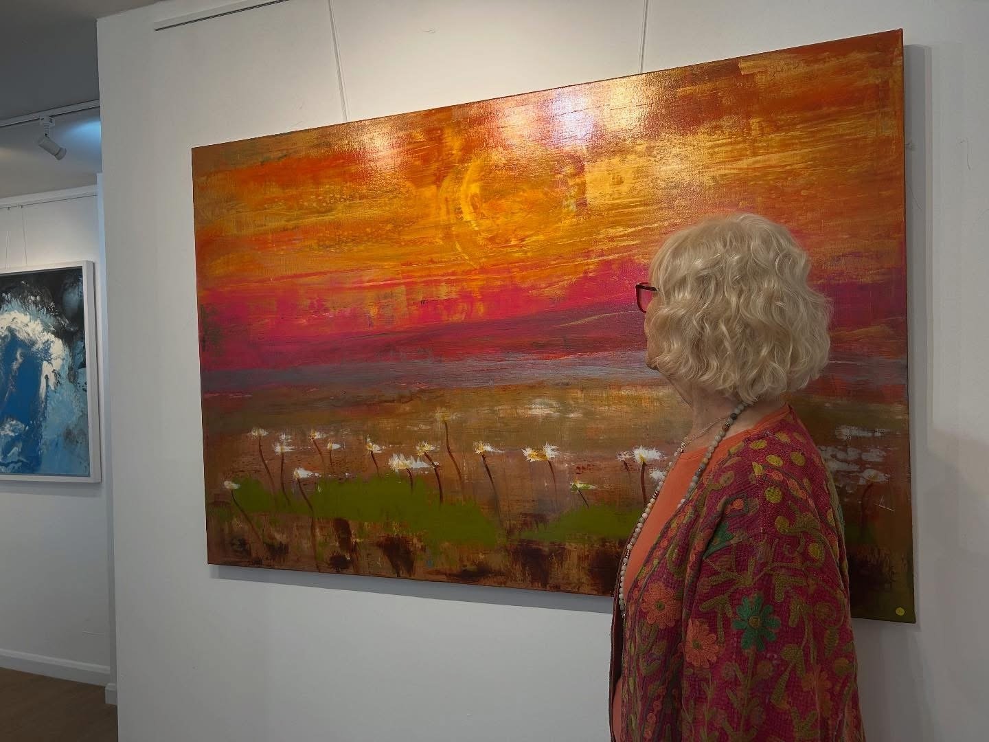 An elderly woman with curly blonde hair and glasses looking at an abstract painting of a sunset over a field with flowers, in an art gallery.