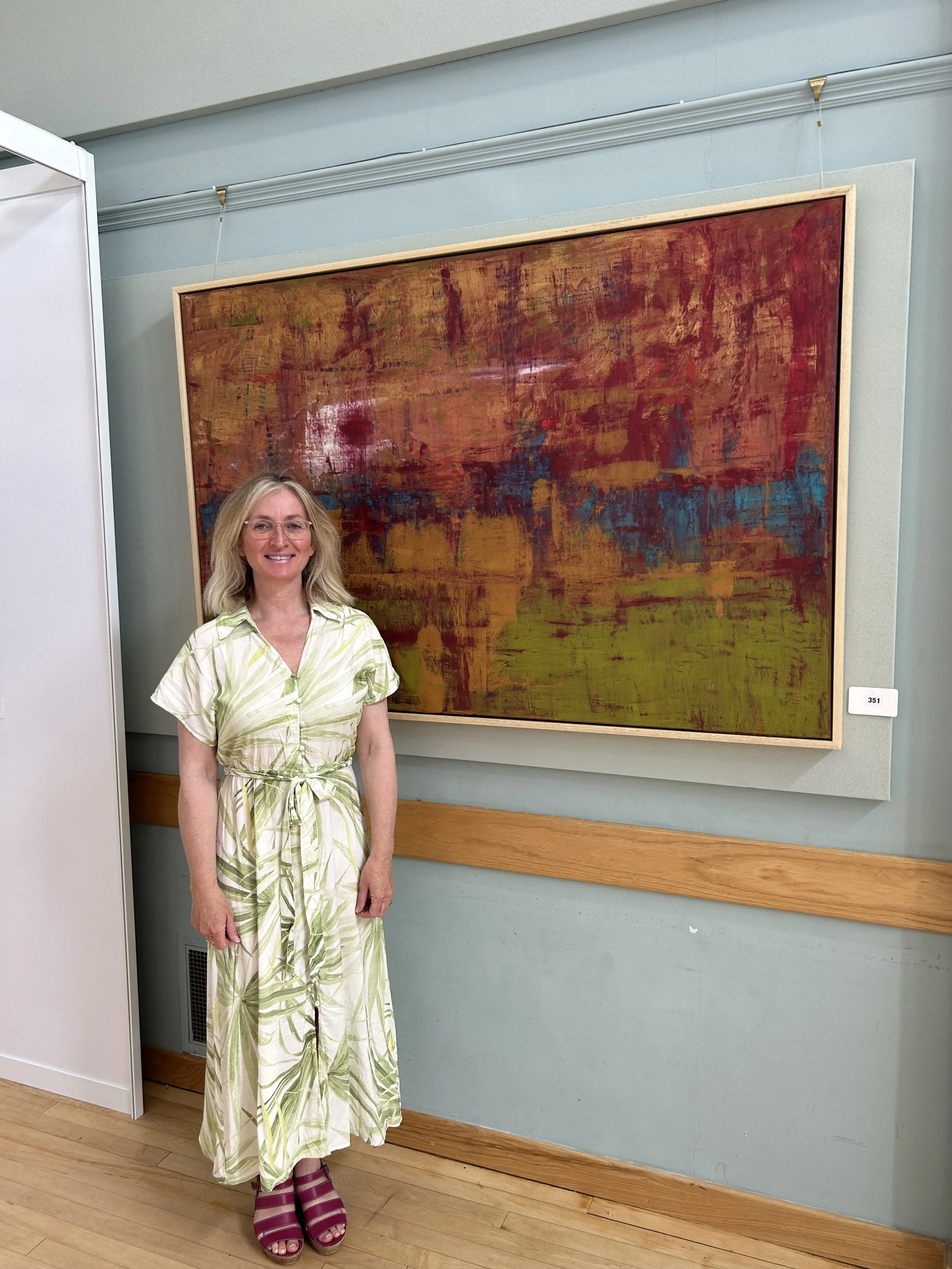 A woman with blonde hair and glasses, wearing a white and green patterned dress and magenta shoes, standing in front of an abstract painting in an art gallery.