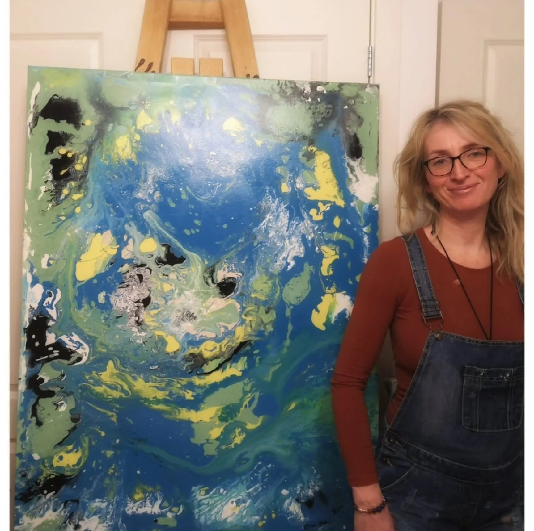A woman with glasses, blonde hair, wearing a rust-colored shirt and denim overalls, standing next to an abstract painting with swirling blue, green, yellow, black, and white colors.