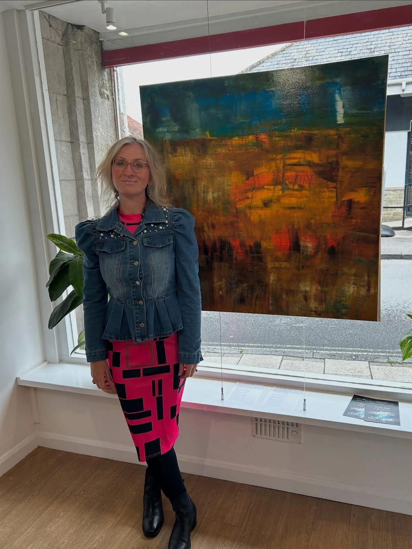 A woman with blonde hair and glasses standing next to an abstract landscape painting in an art gallery, wearing a denim jacket, a pink and black patterned skirt, and black boots.