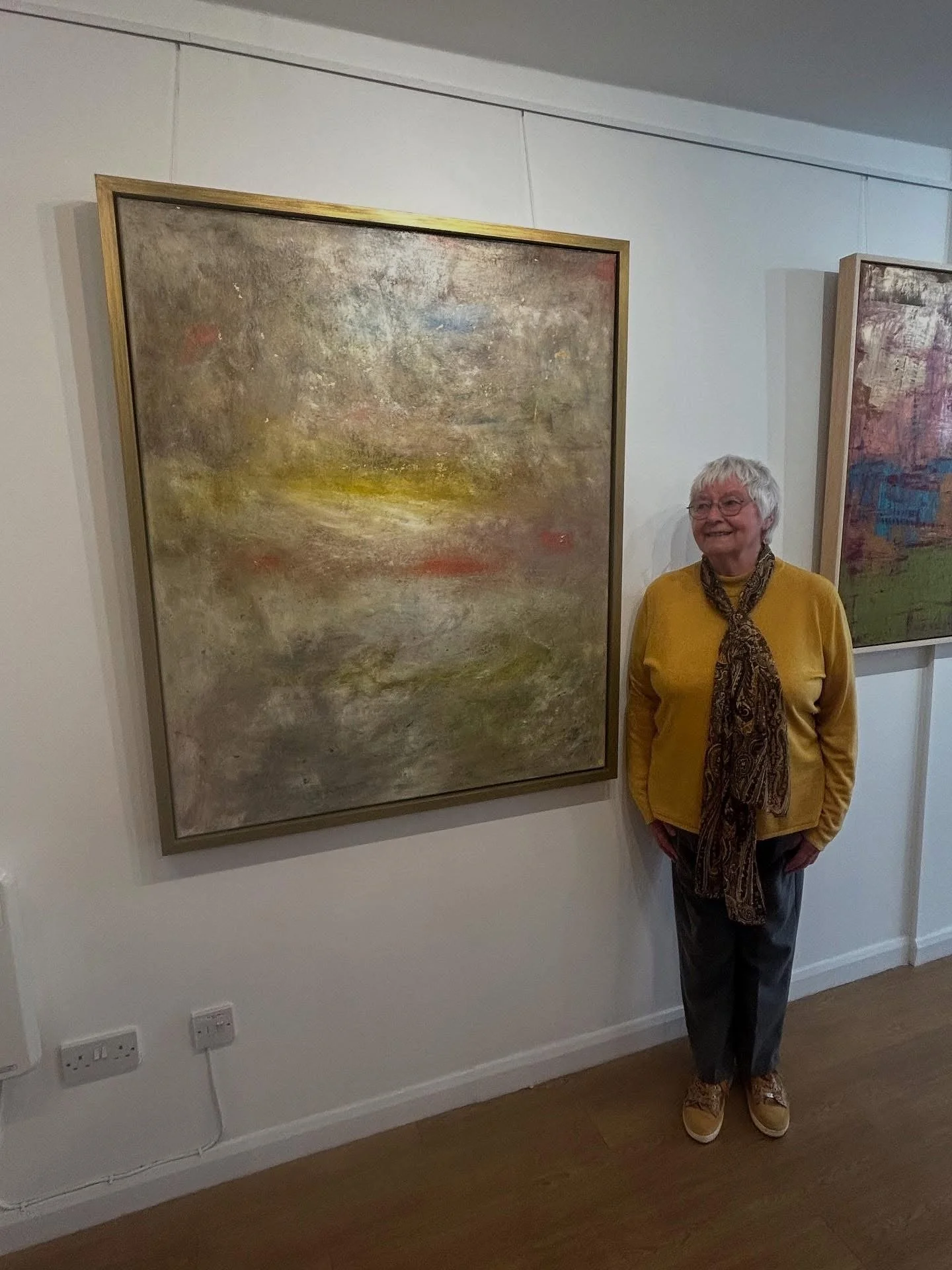 An elderly woman with gray hair, wearing glasses, a yellow sweater, a brown patterned scarf, gray pants, and beige shoes, standing and smiling next to an abstract painting in an art gallery.