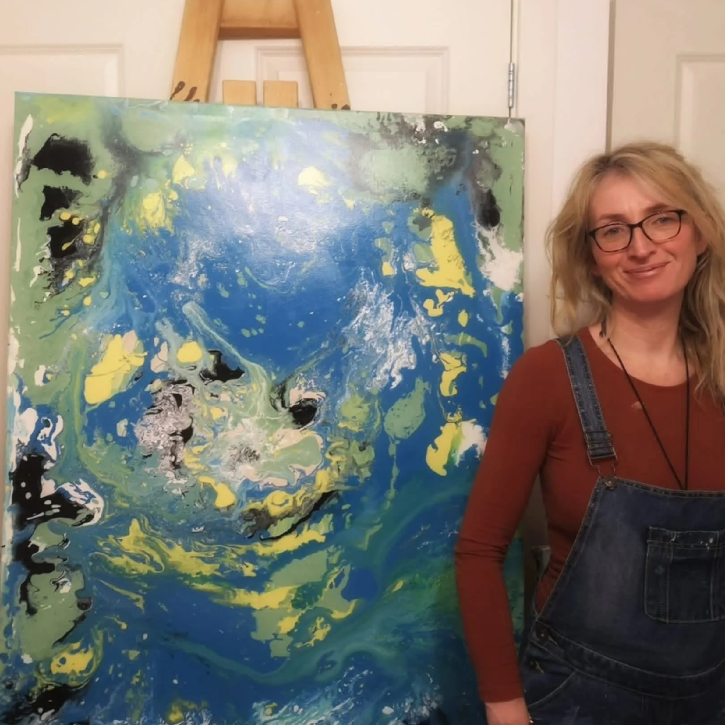 A woman with blonde hair, glasses, wearing a rust-colored top and denim overalls, standing next to an abstract painting with swirls of blue, green, yellow, black, and white.