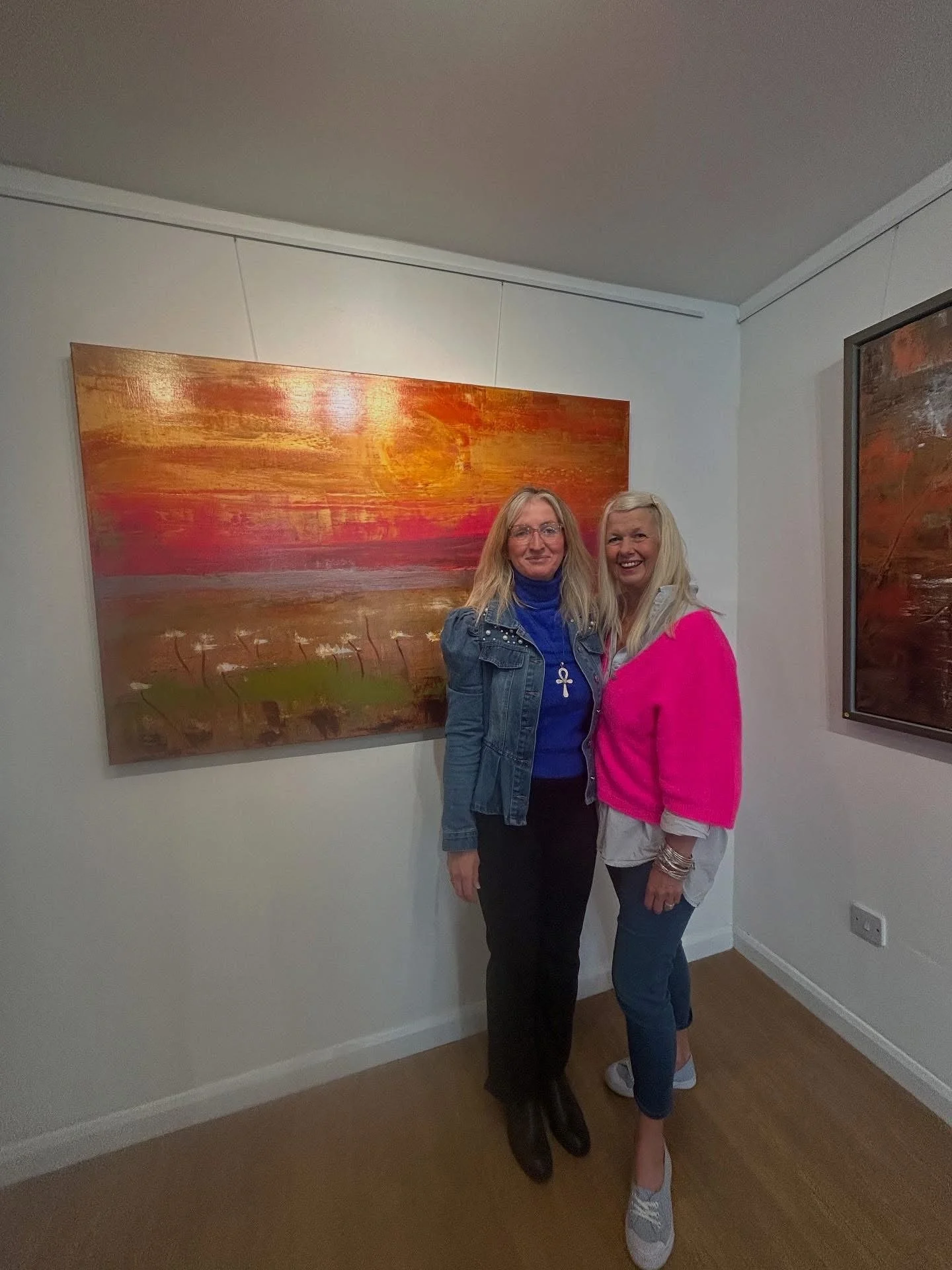 Two women standing in art gallery, posing in front of abstract sunset painting, smiling, one wearing denim jacket and the other in bright pink sweater.