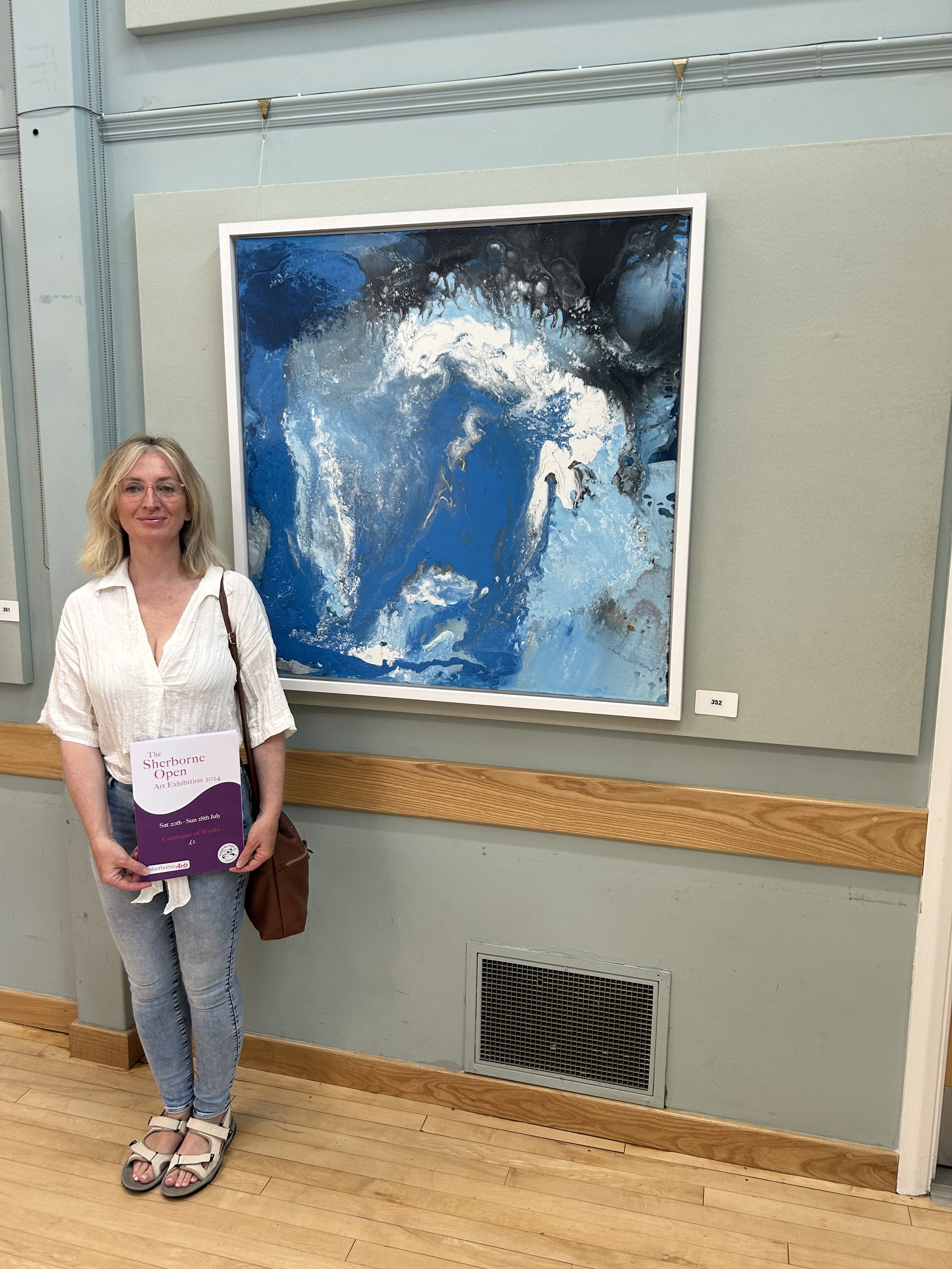 A woman standing in front of an abstract blue and white painting at an art exhibition, holding a brochure for the event.