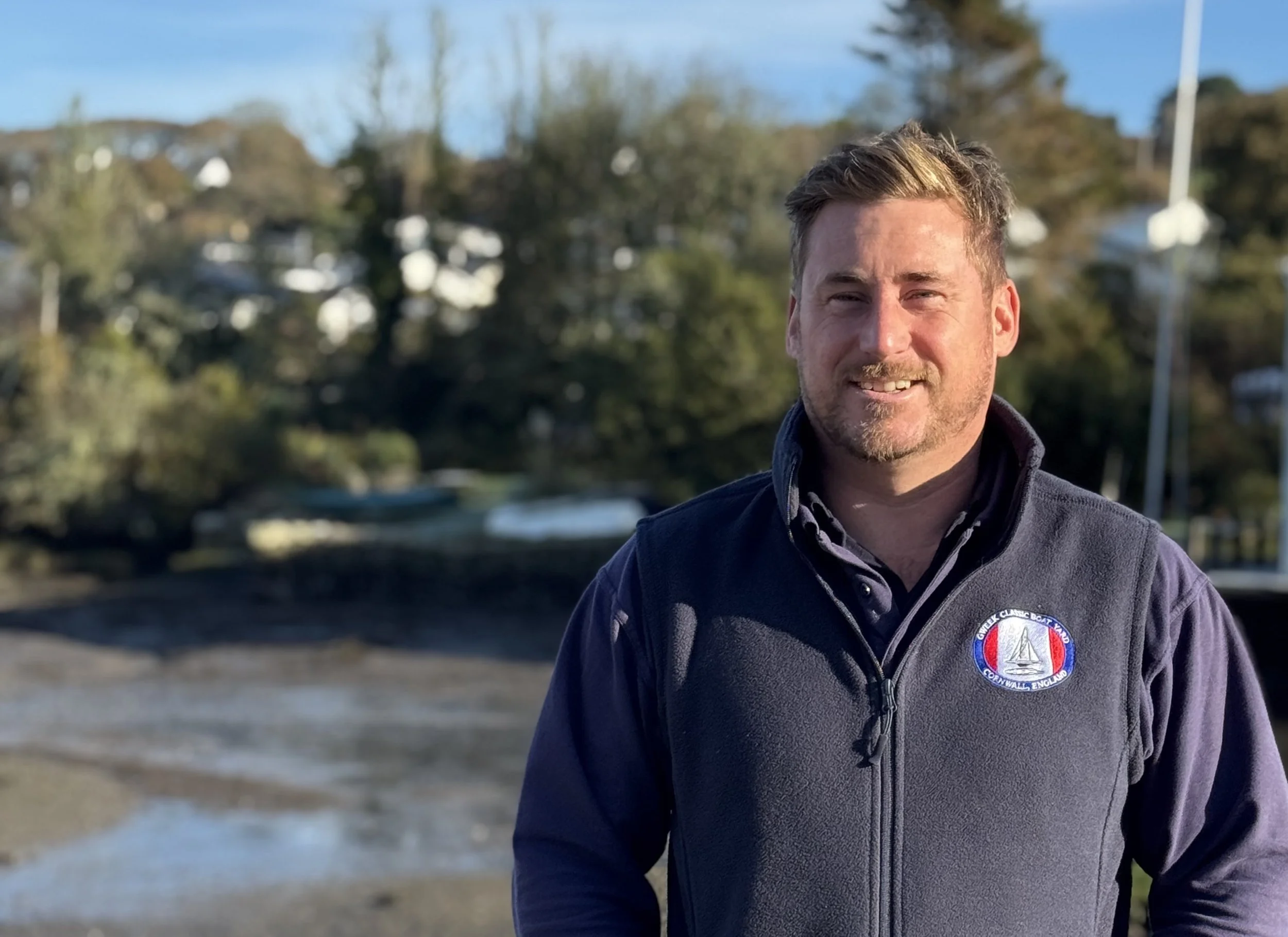 A man standing in front of the river in Gweek wearing a Gweek Classic Boatyard fleece