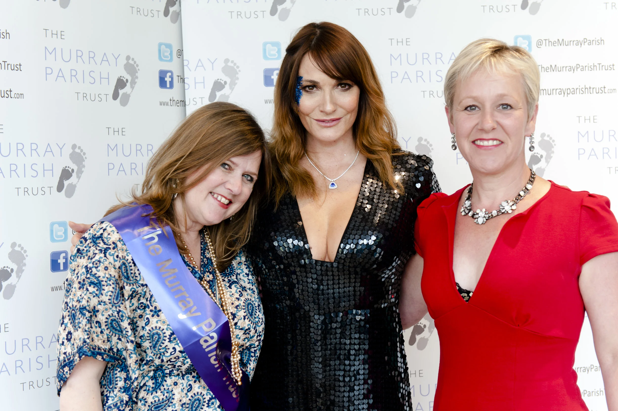 Three women smiling at a formal event in front of a backdrop with the text 'The Murray Parish Trust.' The woman on the left wears a blue and white patterned dress with a blue sash and pearl necklace. The woman in the middle has shoulder-length brown 