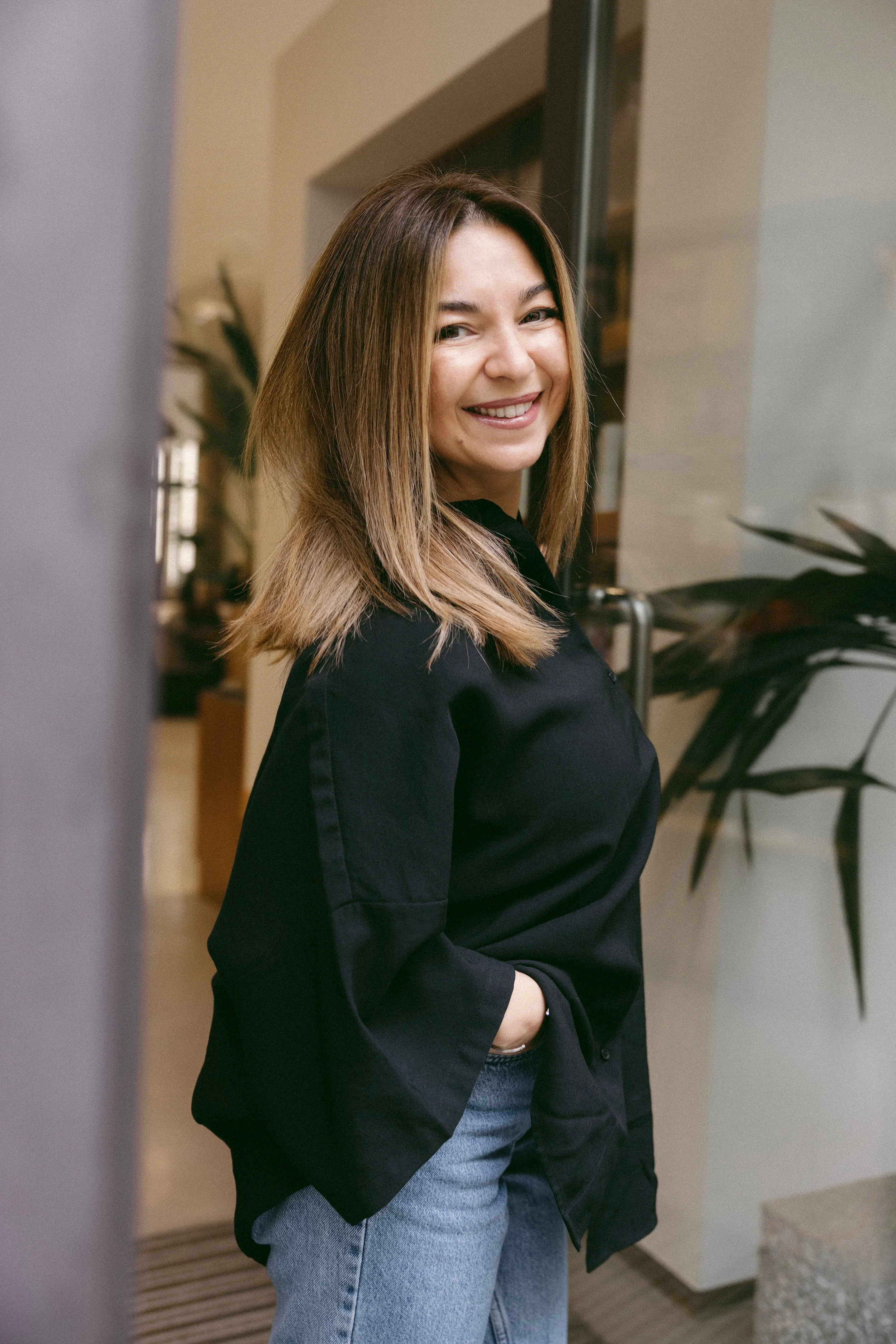 A woman with shoulder-length brown hair, smiling, standing indoors near a wall and plants, wearing a black jacket and blue jeans.