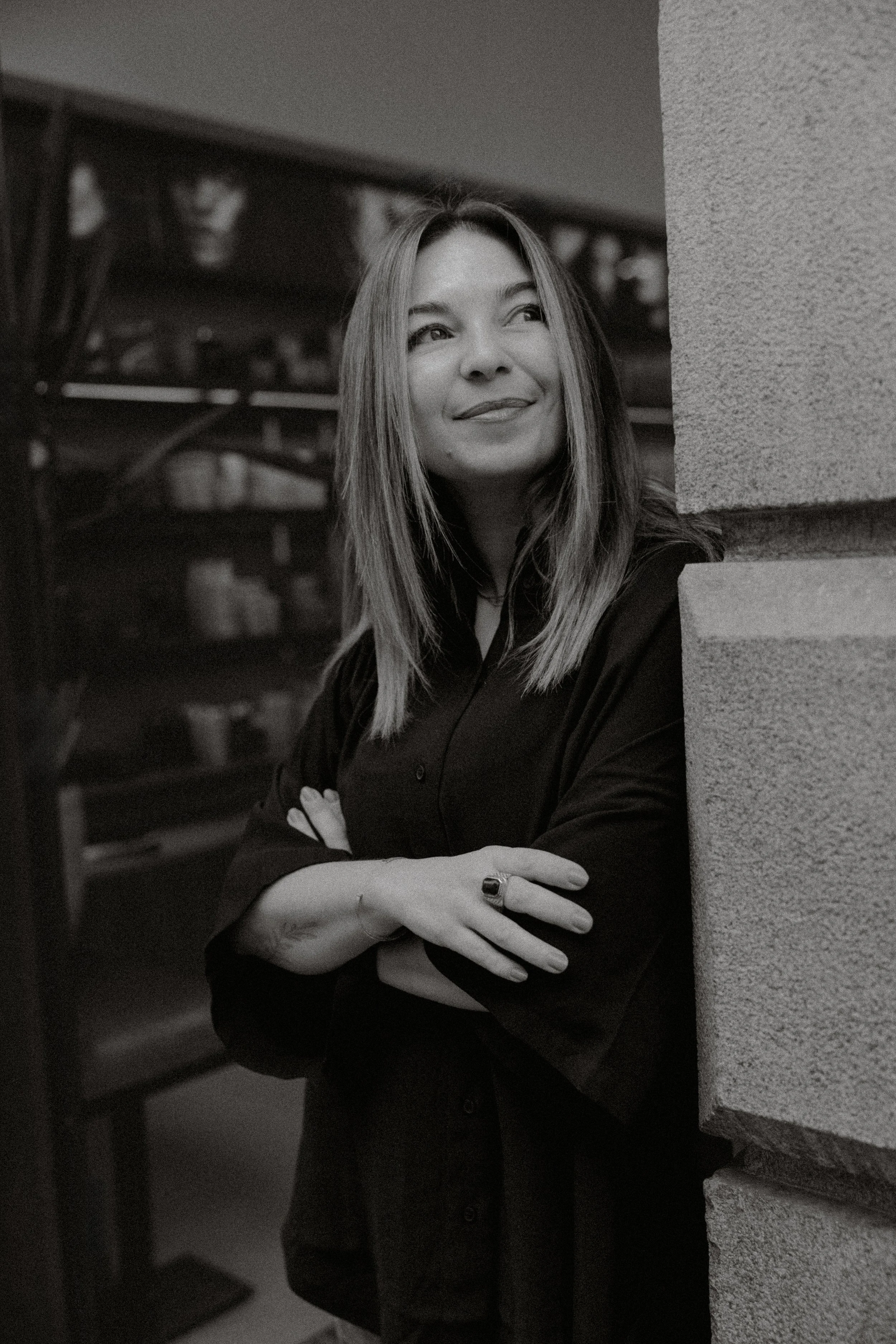 A woman with shoulder-length hair smiling and looking to the side, standing with crossed arms behind a column in an indoor setting with blurred background.