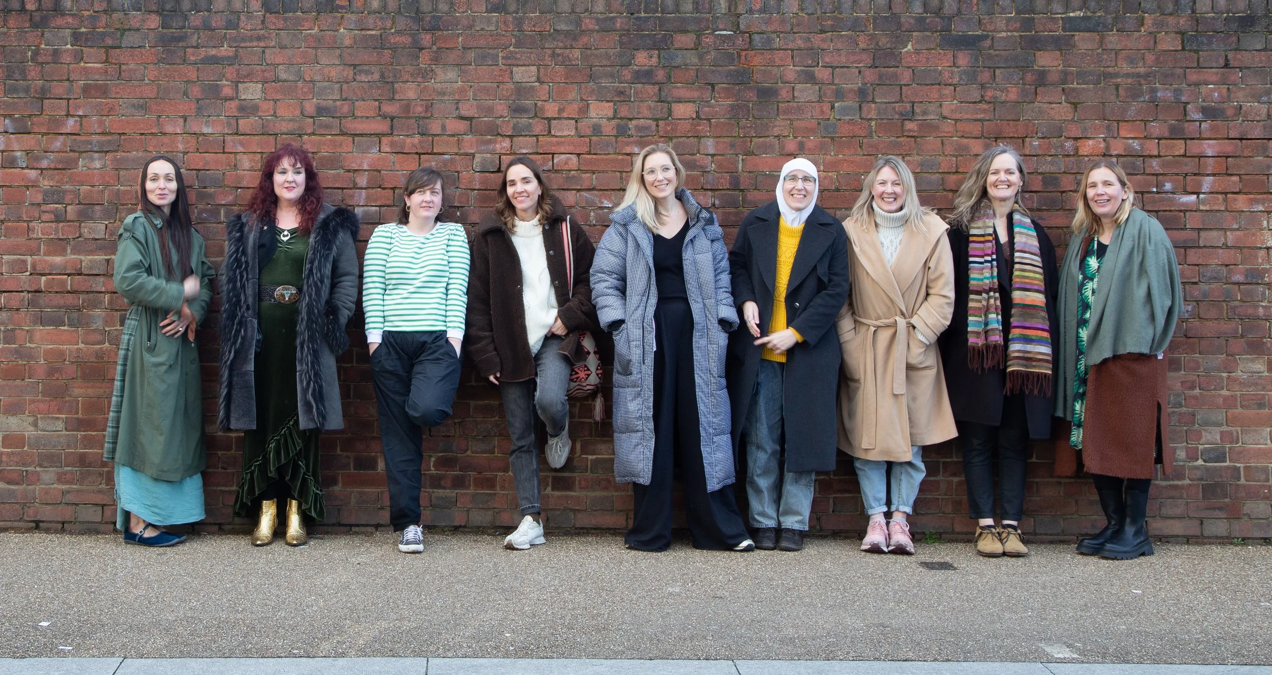 A diverse group of eleven women standing against a brick wall, smiling and posing for a photo, dressed in casual and colorful clothing.