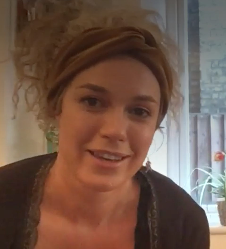 A woman with curly hair and a headband smiling indoors near a window with green plants and potted flowers in the background.