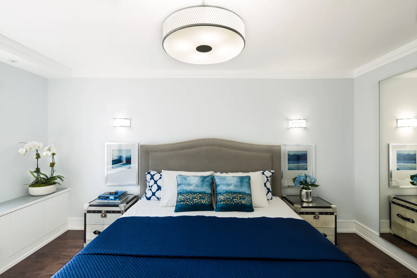 The master bedroom color palette is neutral gray and white accented with blue bedding and artwork, very easy to swap if desired. A full length mirror bounces off the natural light from the windows on the opposite wall. This bedroom is a textural dream with the cowhide on the nightstands, leather and nail head trim, velvet headboard, quilted bedding and the softness from the flowers! Planned layers are critical to making a room come together. We loved designing and styling this room. 