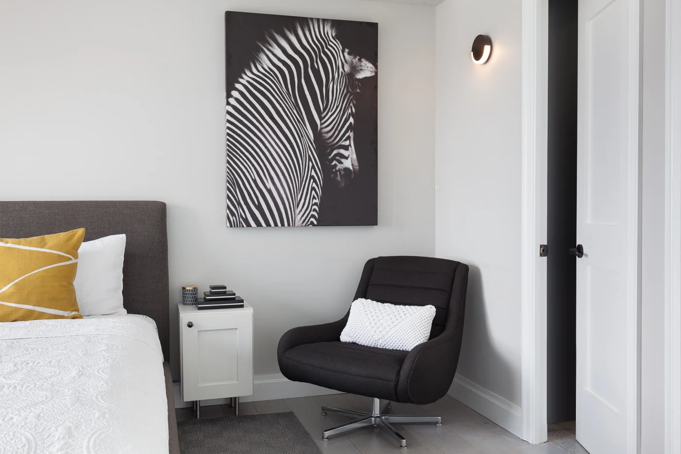 Keeping with the black and dark moody greys throughout the house, we bring in the black and white zebra art. The art pulls together the room with the dark grey swivel chair making it the perfect spot for that morning cuppa joe. I love the slight peek into the custom dark wood walk in closet and the minimal black sconce we selected for lighting here. The crisp white quilt with the bright chartreuse is the perfect pop of color without being too obvious.