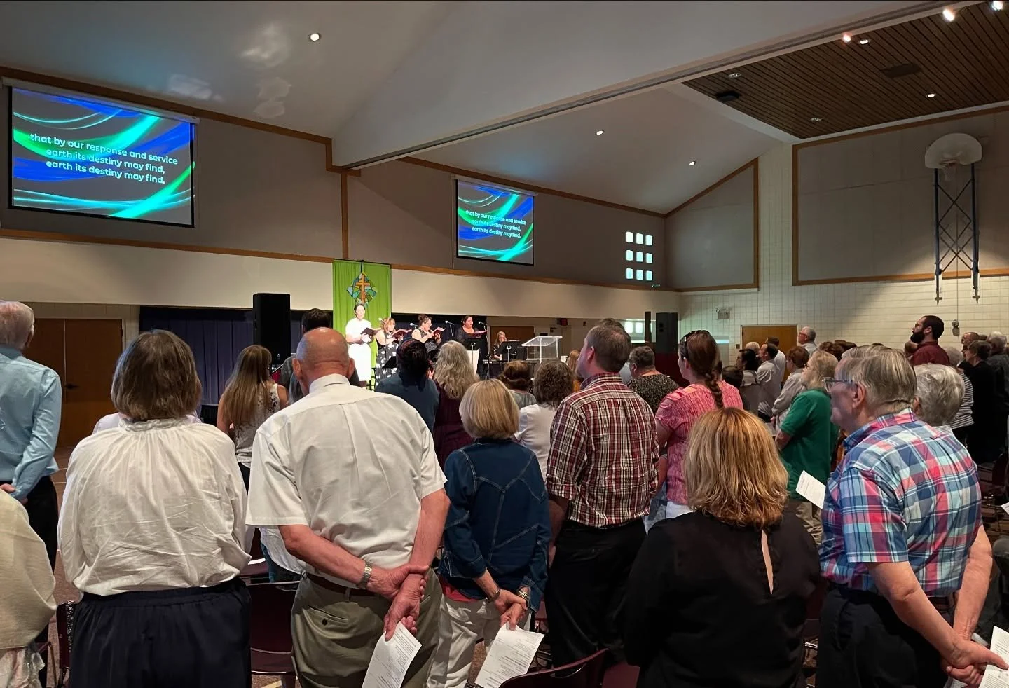 A congregational gathering in a church with people standing and listening to a choir on stage, with lyrics displayed on screens: 'earth by our response and service, earth its destiny may find.'