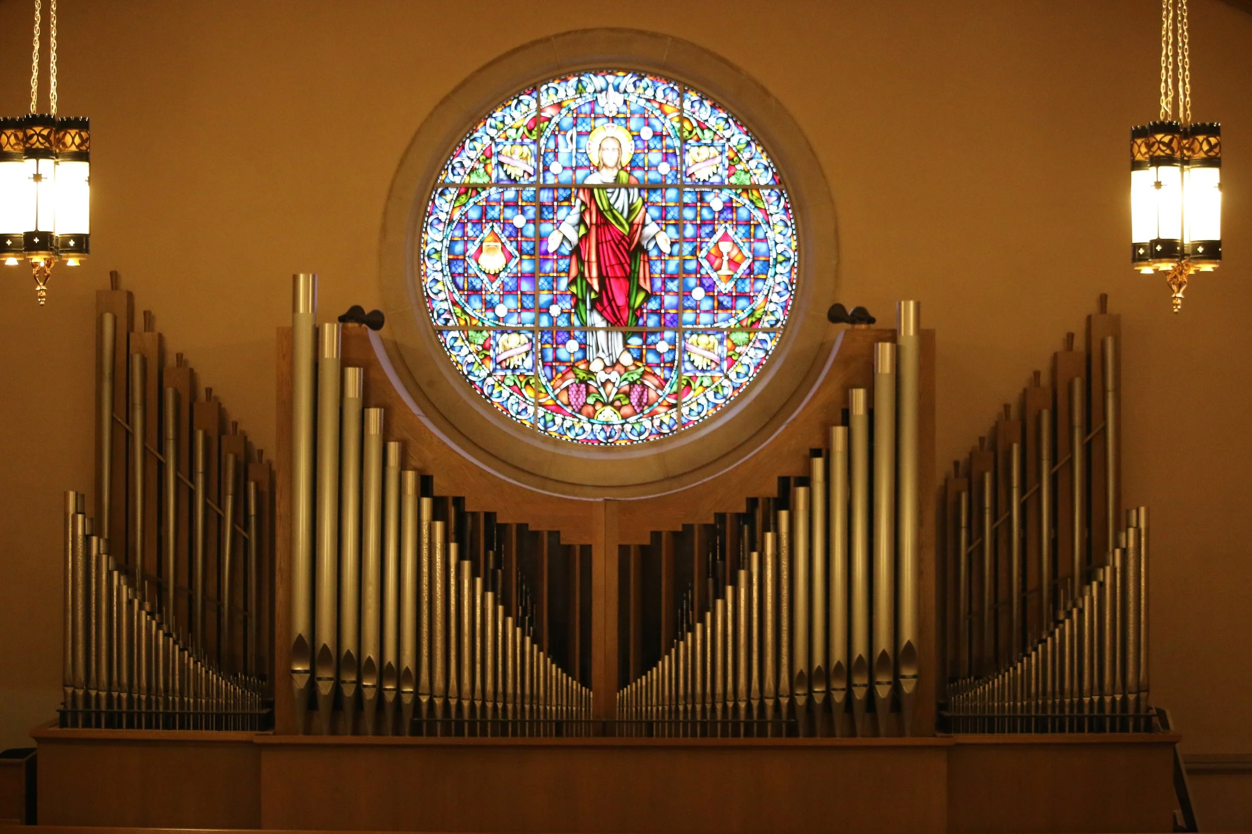 Stained glass window depicting Jesus Christ in a church, above an organ with multiple pipes, illuminated by two hanging lamps.