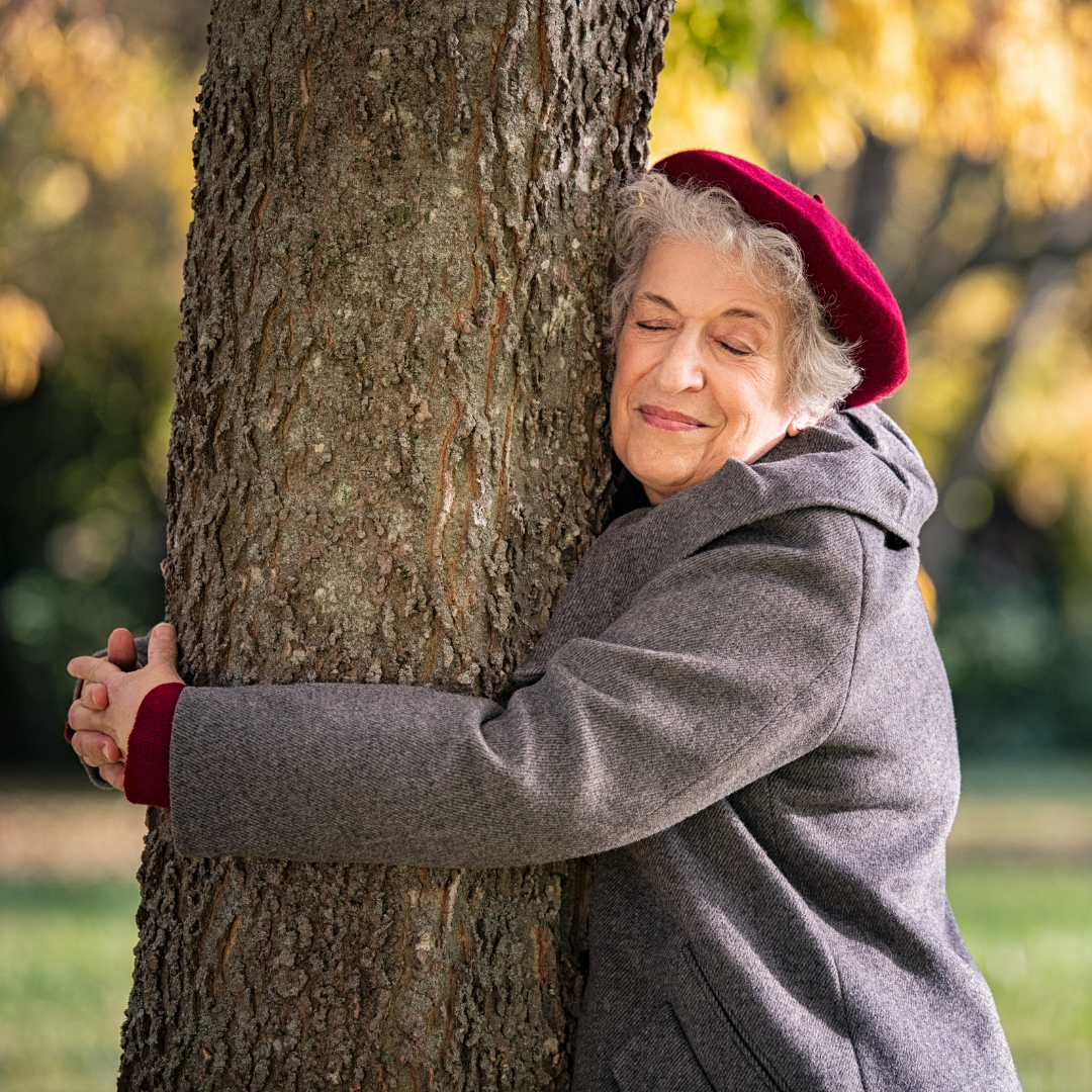 An older woman wearing a beret smiles as she embraces a tree, symbolizing joy and connection with nature.