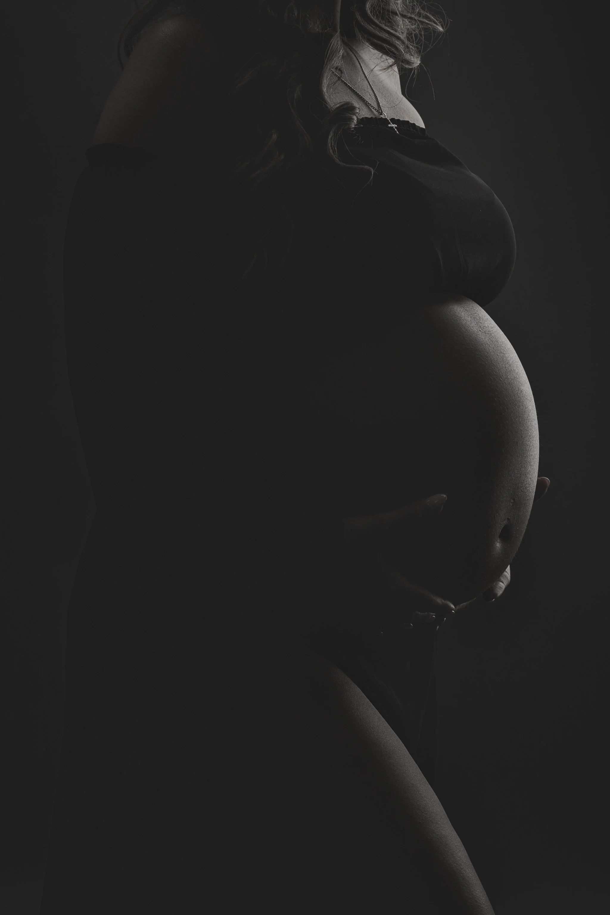 Artistic black and white maternity portrait highlighting the baby bump with soft shadows and wisps of hair, photographed at Fotoplicity studio in East Brunswick NJ