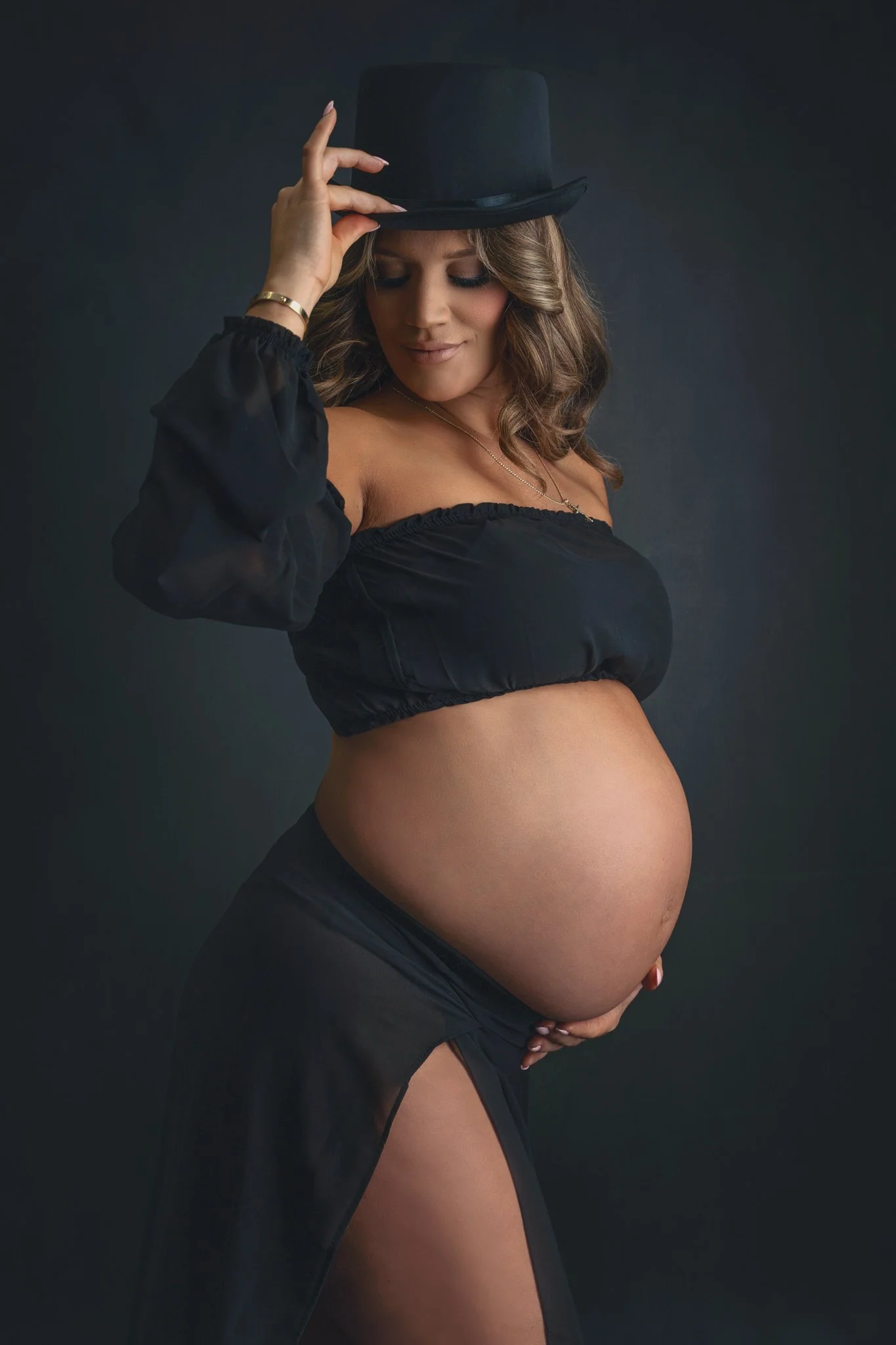 Maternity portrait of a woman in a halter top and slitted skirt wearing a top hat, showcasing her baby bump on a dark gray backdrop at Fotoplicity studio in East Brunswick NJ