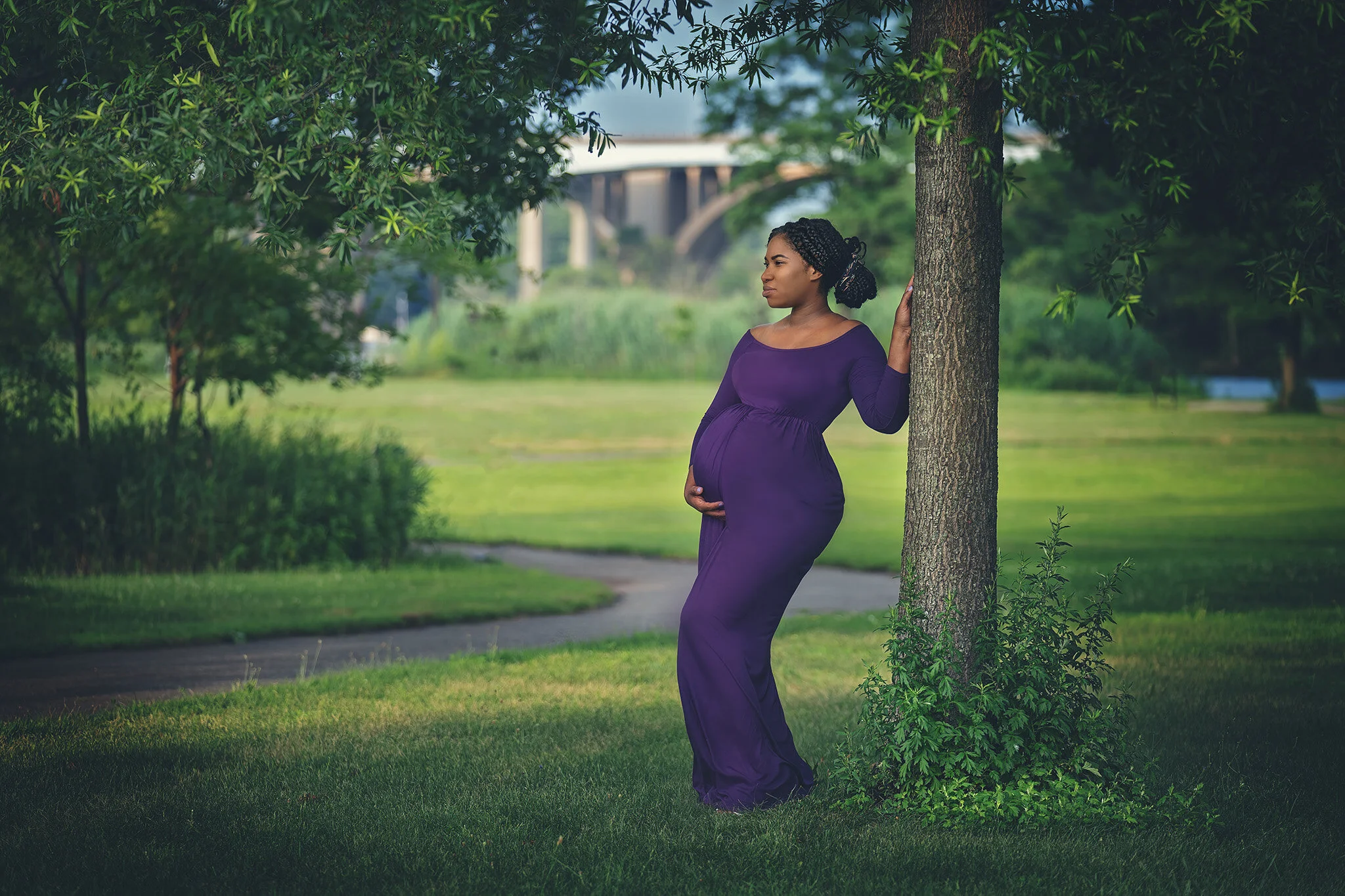 Pregnant woman in a purple dress leaning against a tree during a maternity photo session at Donaldson Park in Highland Park, New Jersey, with the Raritan River bridge in the background.