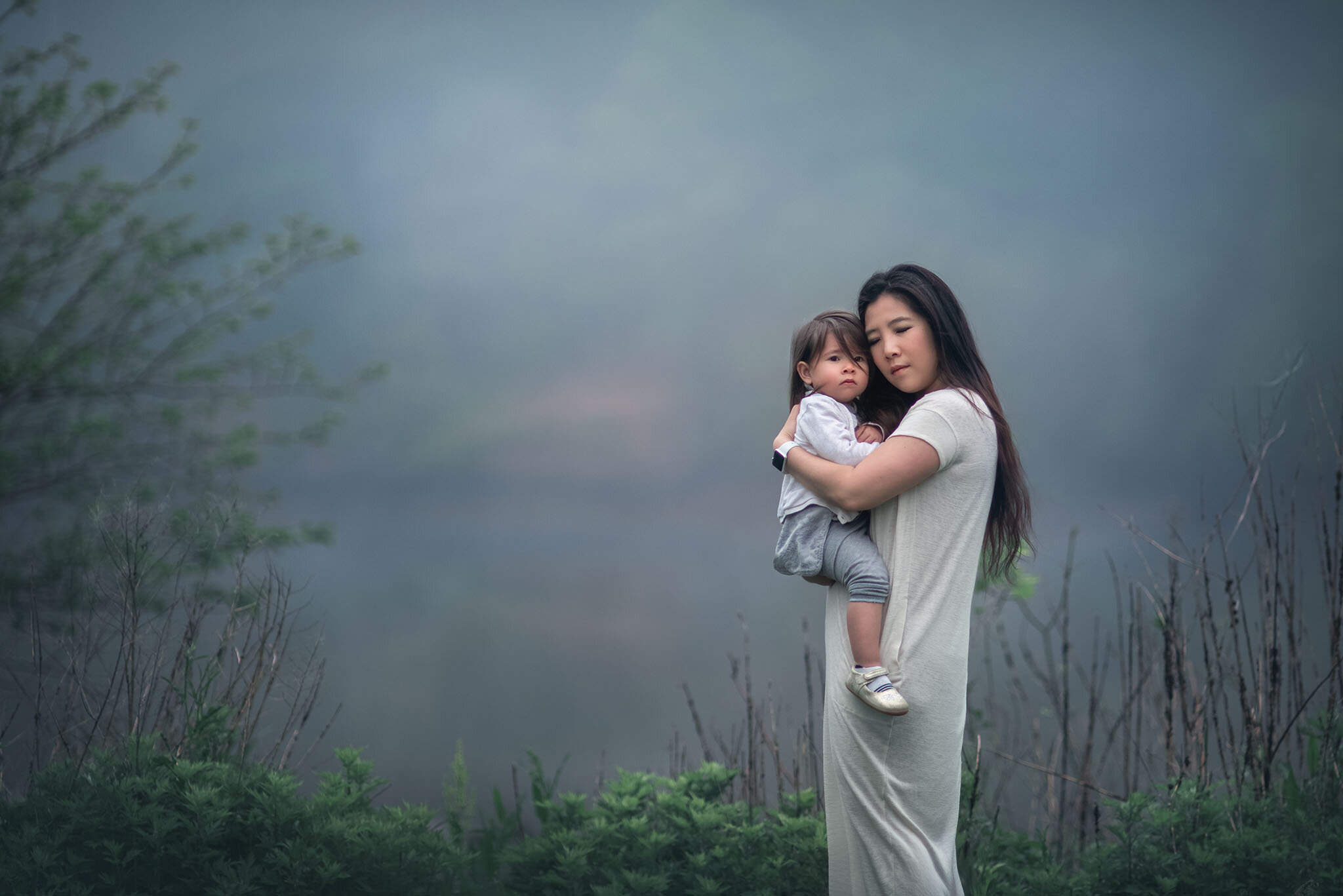 Loving outdoor family portrait of a mother holding her child against her chest with eyes closed, overlooking the foggy Raritan River at Donaldson Park in Highland Park NJ, photographed by Fotoplicity