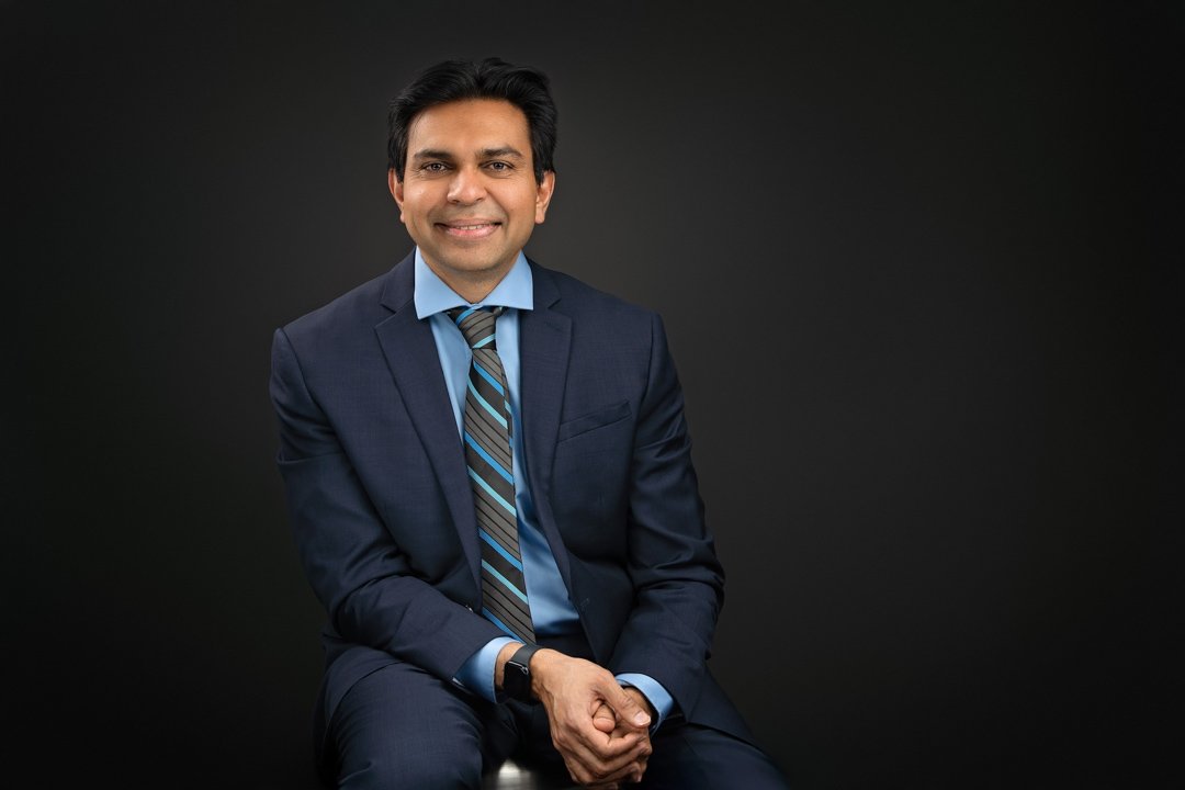 A professional man sitting on a chair for a headshot with a dark gray backdrop.