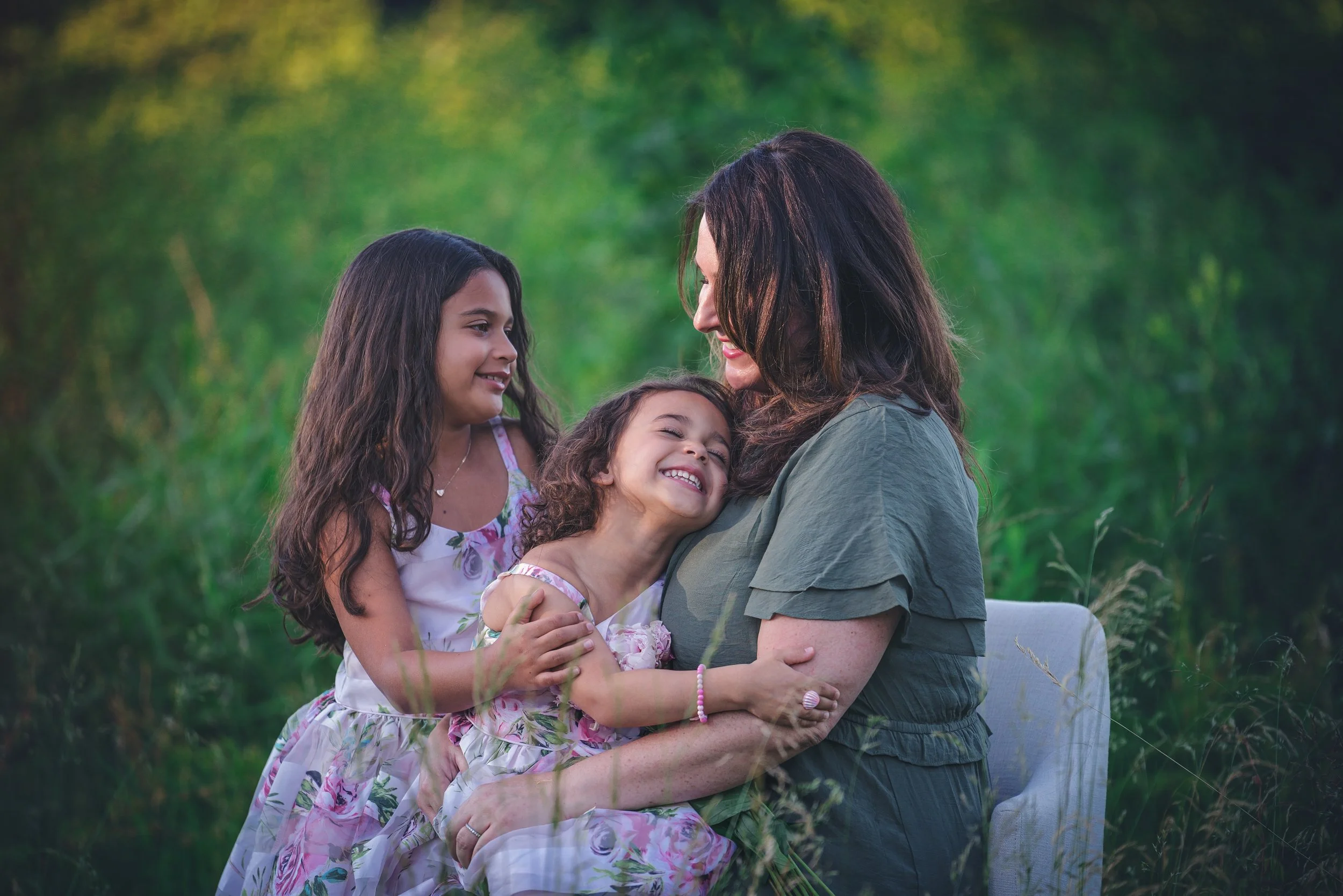 Mother snuggling her two young children in a tall green field during a natural light outdoor family photography session in New Jersey.