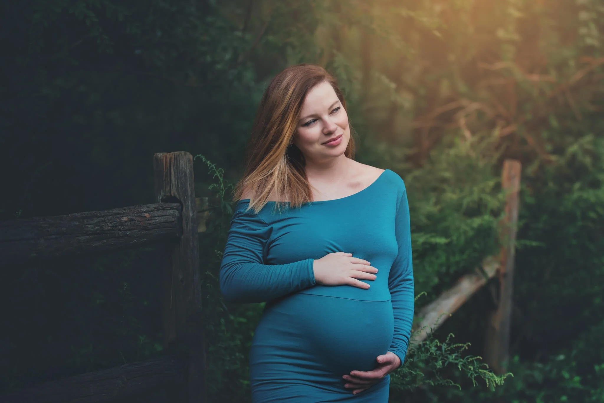Pregnant woman in a teal dress cradling her baby bump during an outdoor maternity portrait session with Fotoplicity in New Jersey
