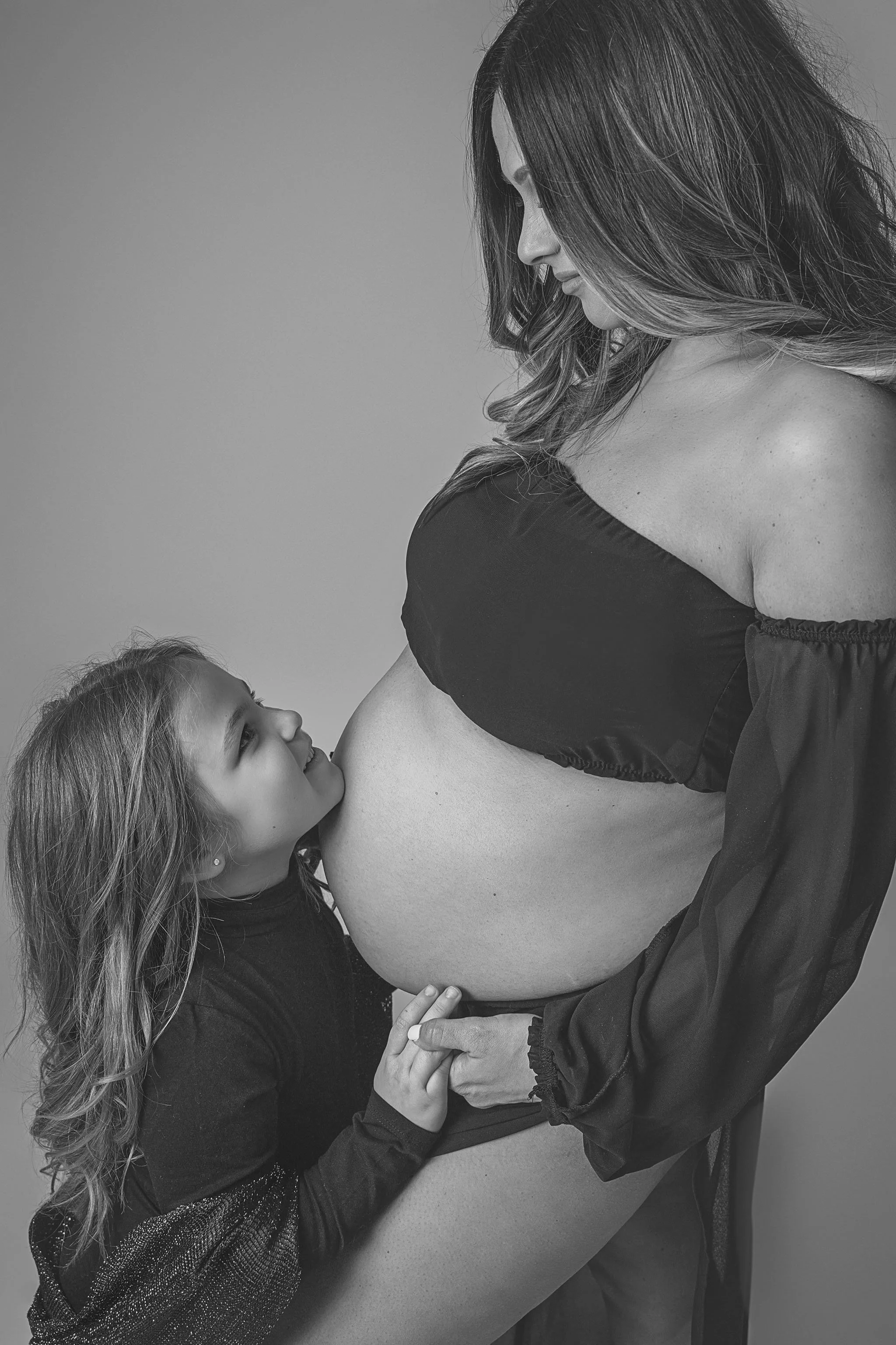 Loving maternity portrait of a mother and young daughter holding hands with daughter's chin resting on mom's belly, soon to be siblings, at Fotoplicity studio in East Brunswick NJ