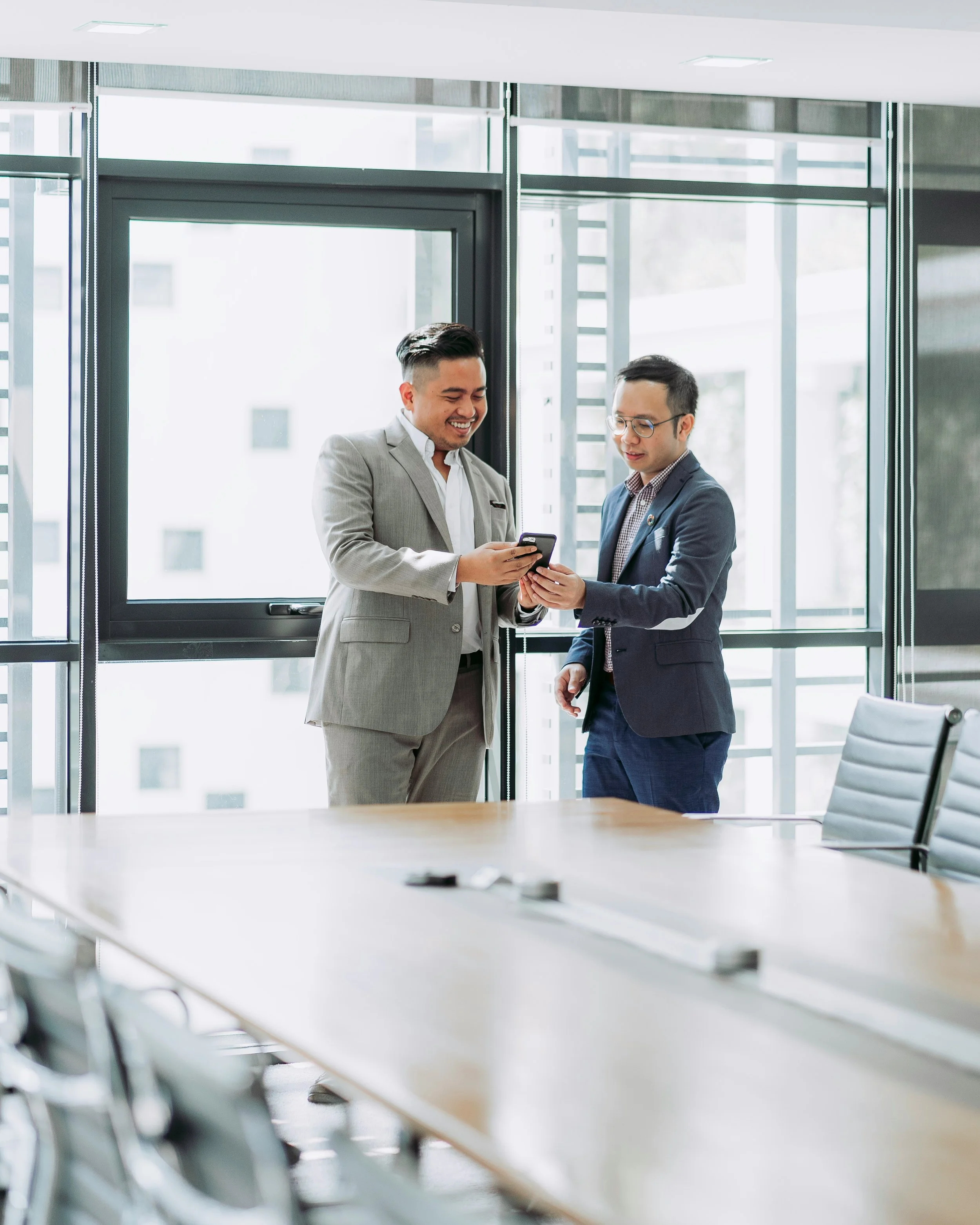 Two men in business suits standing in an office, looking at a smartphone and smiling.