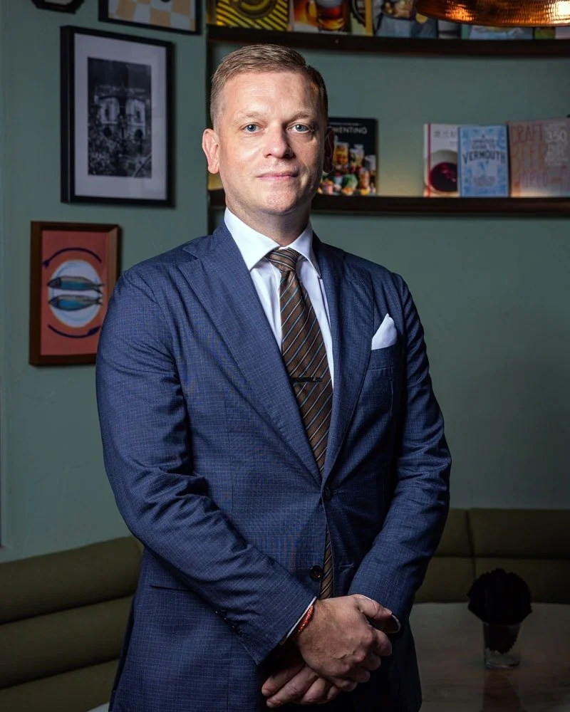 A man in a blue suit and striped tie standing with arms crossed in a room decorated with framed pictures and books on a shelf.