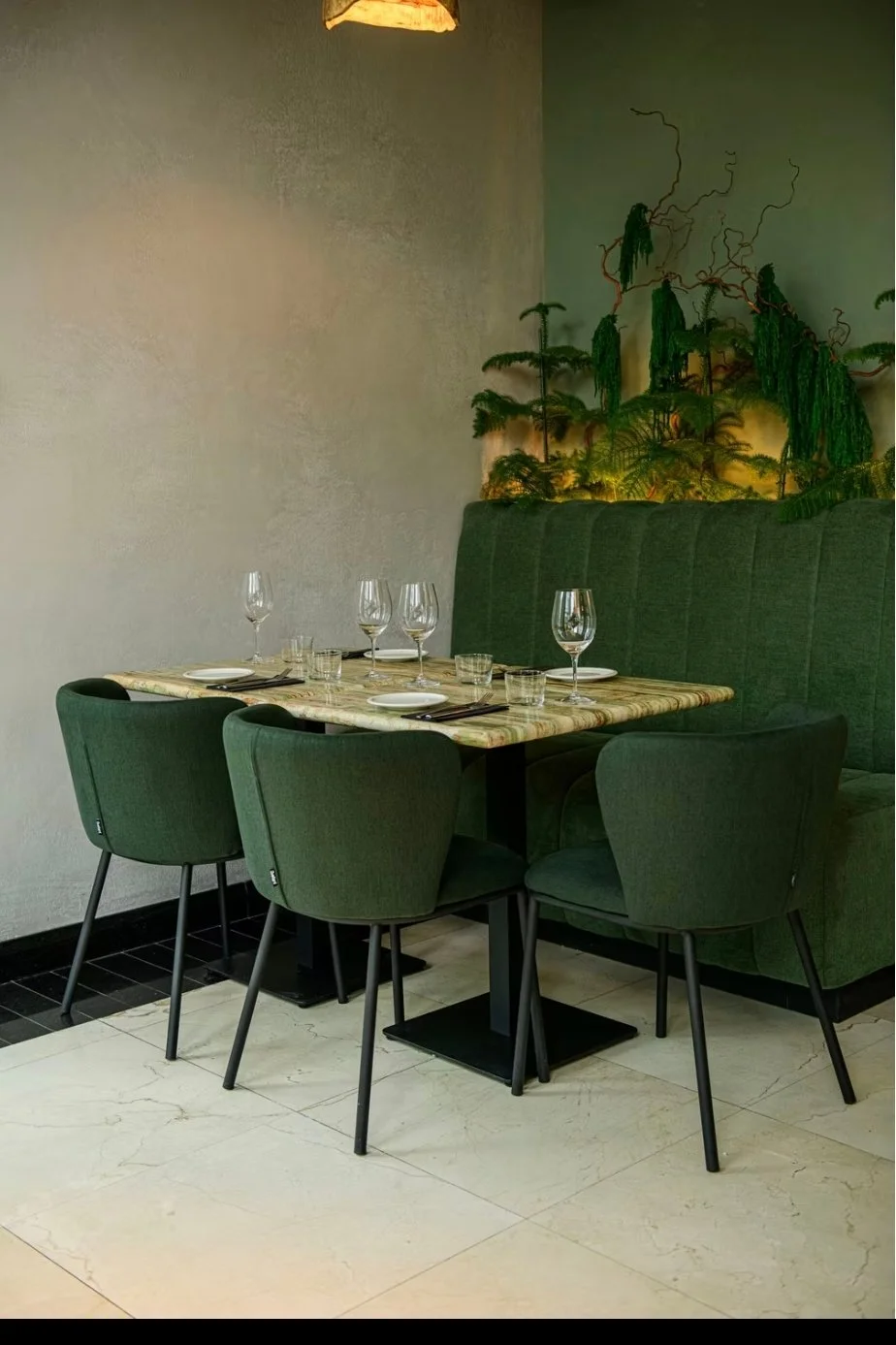 A dining table set with wine glasses, plates, and cutlery in a restaurant with green chairs and a green upholstered banquette, decorated with a leafy green wall art installation.