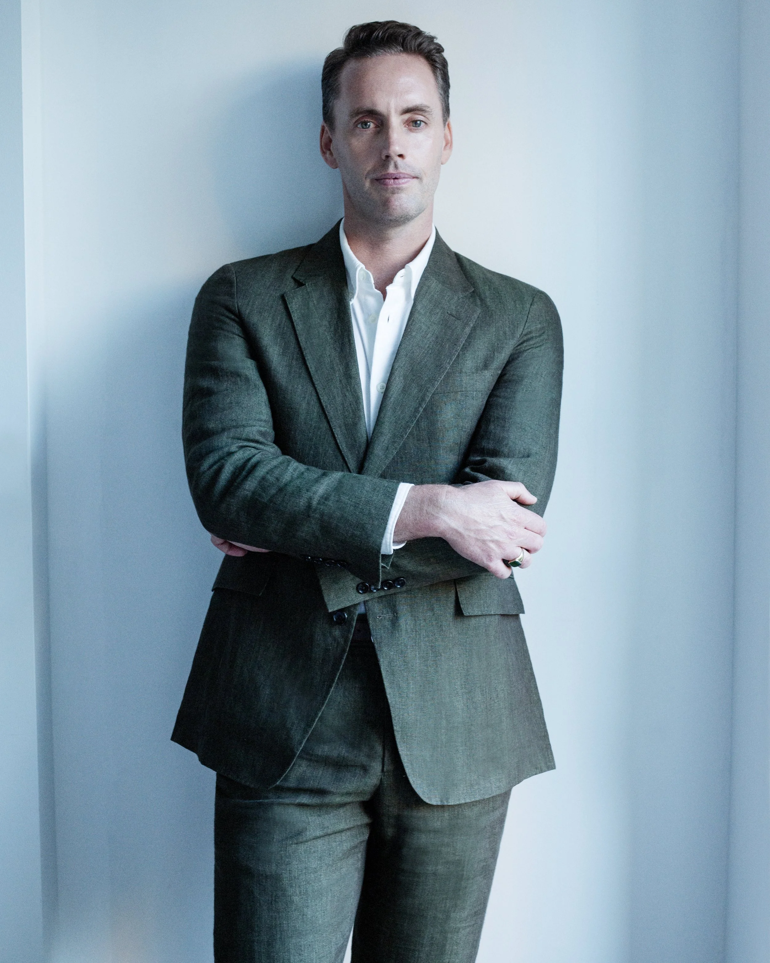 A smiling man with short hair, wearing a dark green suit and white shirt, standing against a light blue wall.