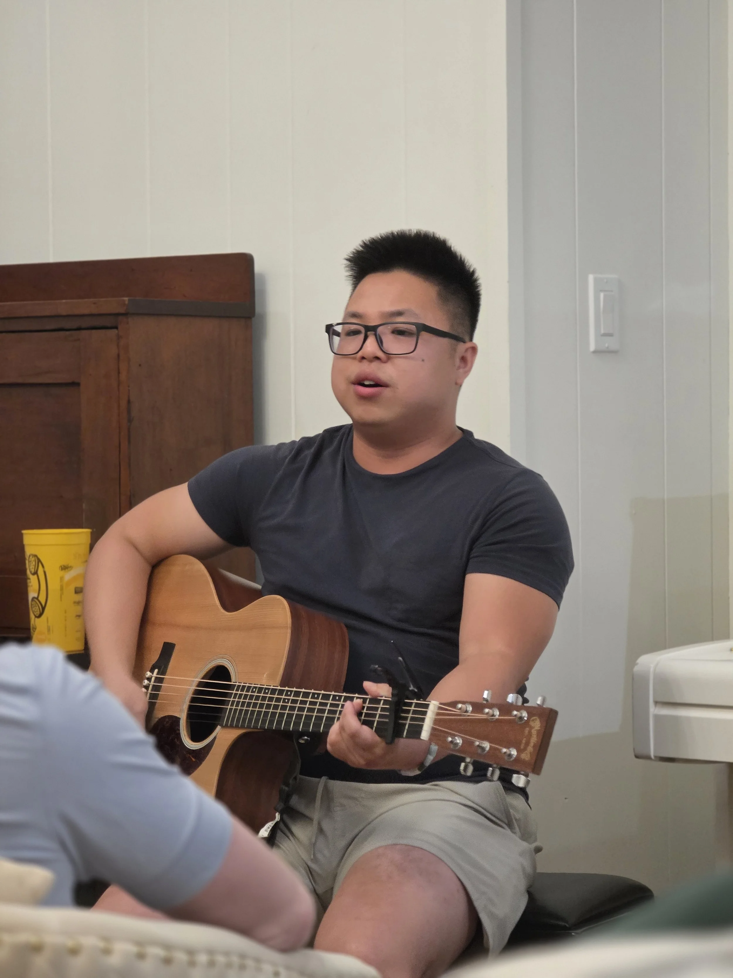 An asian man sitting playing an acoustic guitar.