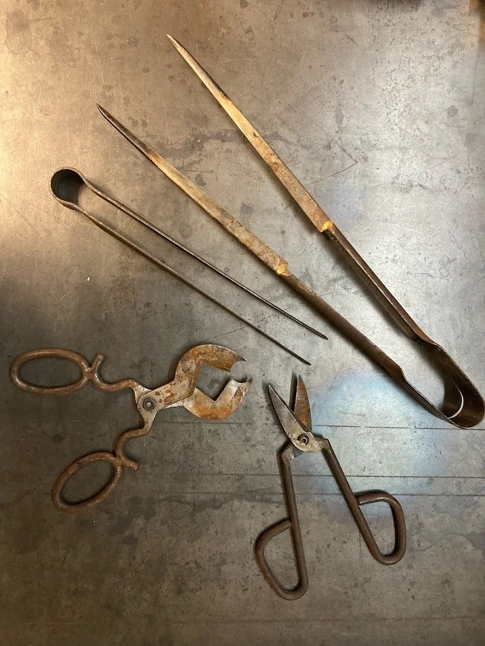 A collection of vintage metal tools, including two large, rusty tweezers with long, pointed tips, a pair of small, rusty shears with finger loops, and a curved metal hook.