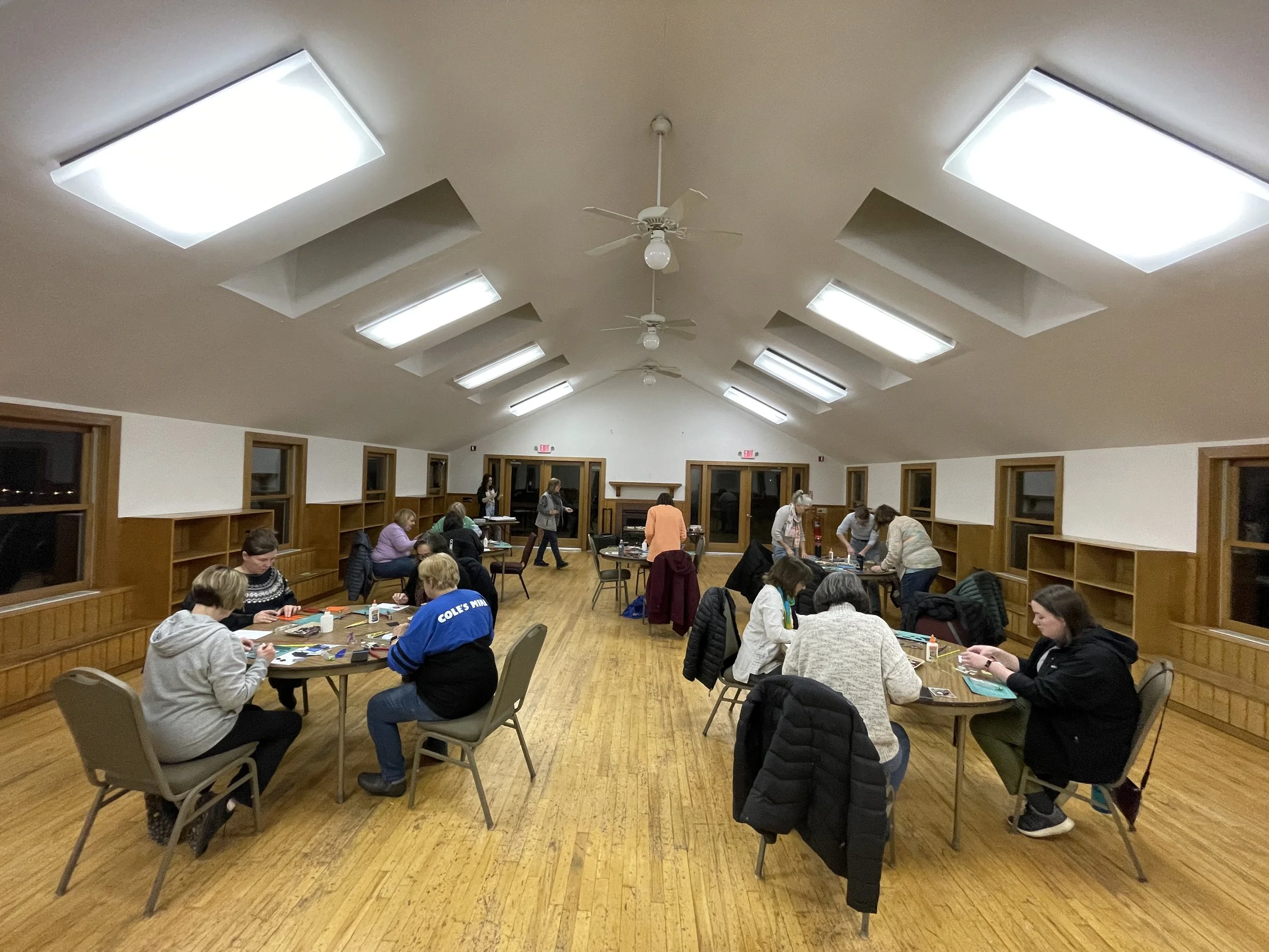 People participating in a craft workshop in a large, well-lit room with wooden floors and built-in wooden shelves along the walls.
