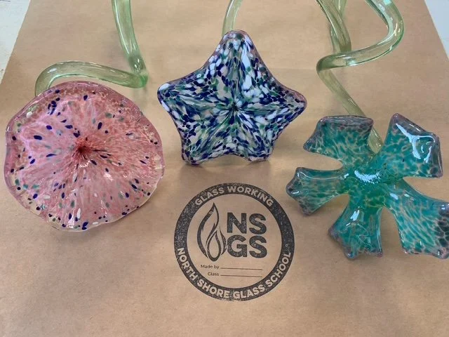 Three colorful glass star-shaped art pieces on a wooden surface, with a background of industrial kitchen equipment and utensils.