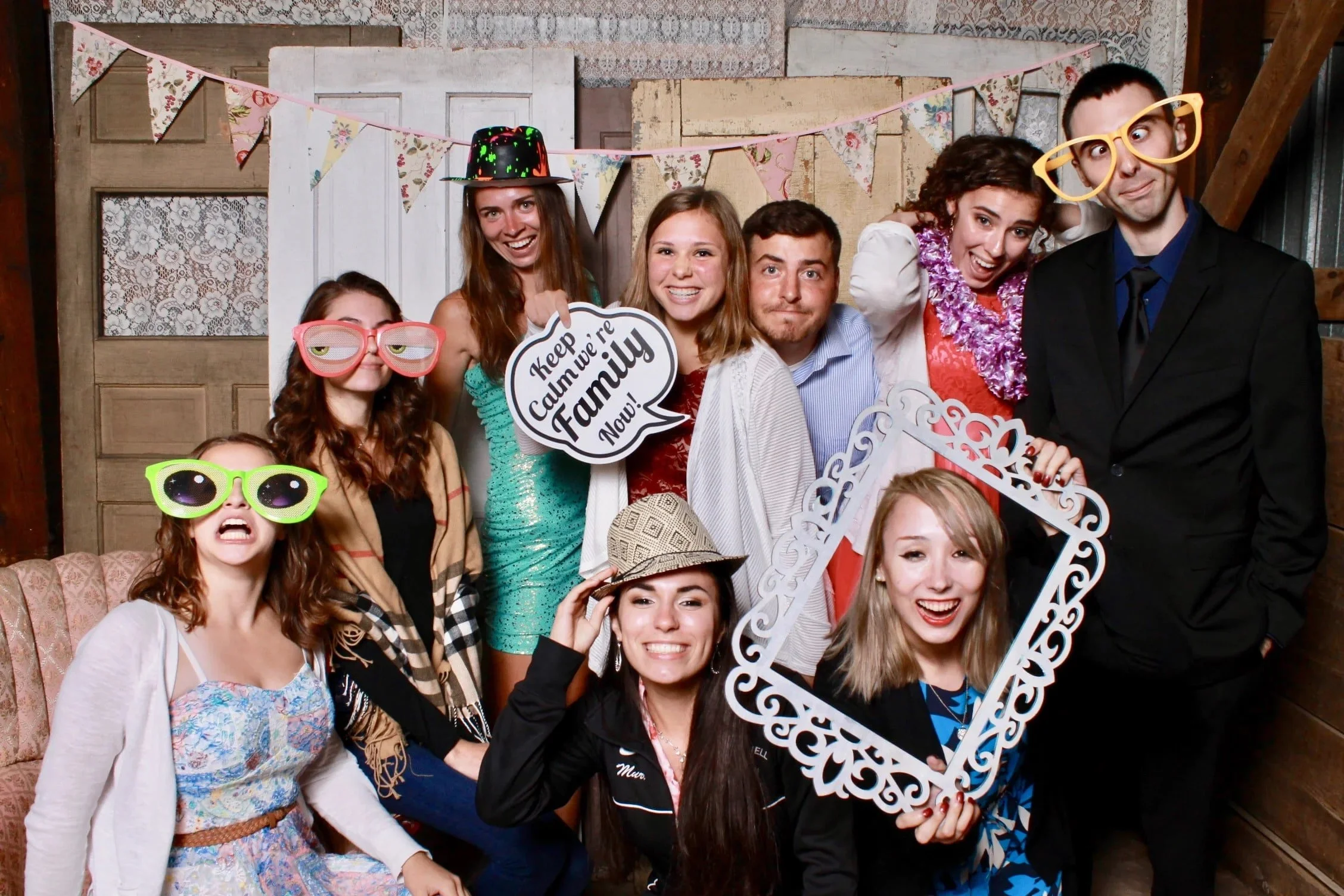 Group of ten young adults at a party, wearing fun glasses and hats, holding a decorative frame and a speech bubble sign, in a rustic decorated room with bunting and lace curtains.