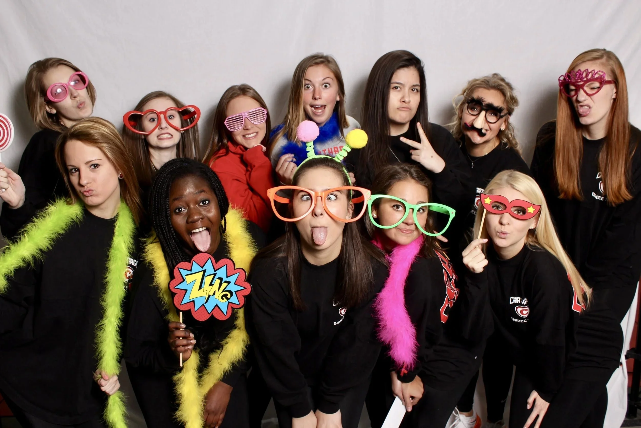 Group of young women wearing humorous glasses, colorful feather boas, and holding fun props in a playful pose