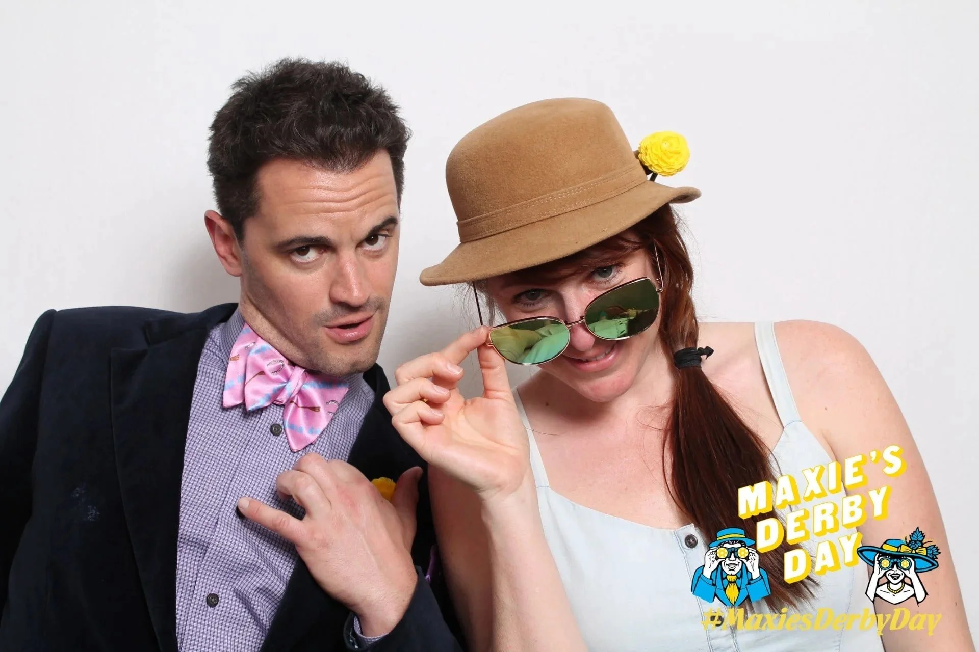 A man and woman posing playfully against a plain white background. The man is wearing a black blazer, a checkered shirt, and a pink bow tie with a pattern, looking at the camera with a surprised expression. The woman is wearing a tan hat with a yellow flower, sunglasses, and a white sleeveless top, adjusting her sunglasses and looking at the camera. Text on the image reads "Maxie's Derby Day" with a hashtag #MaxiesDerbyDay and cartoon illustrations of people with hats.