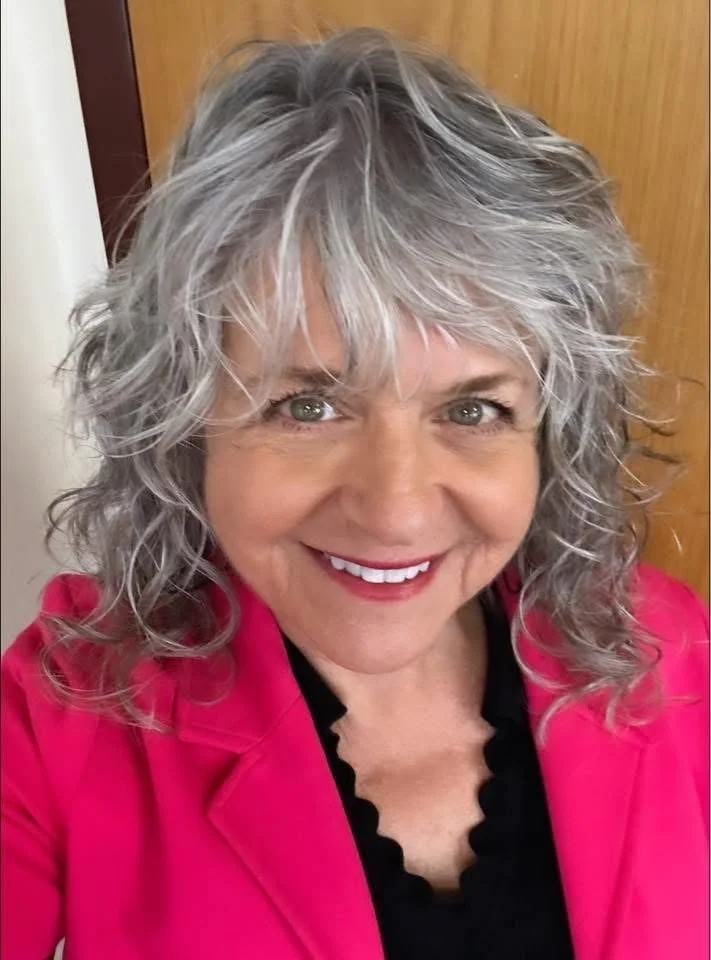 A woman in a pink dress jacket, black blouse, with greyish hair