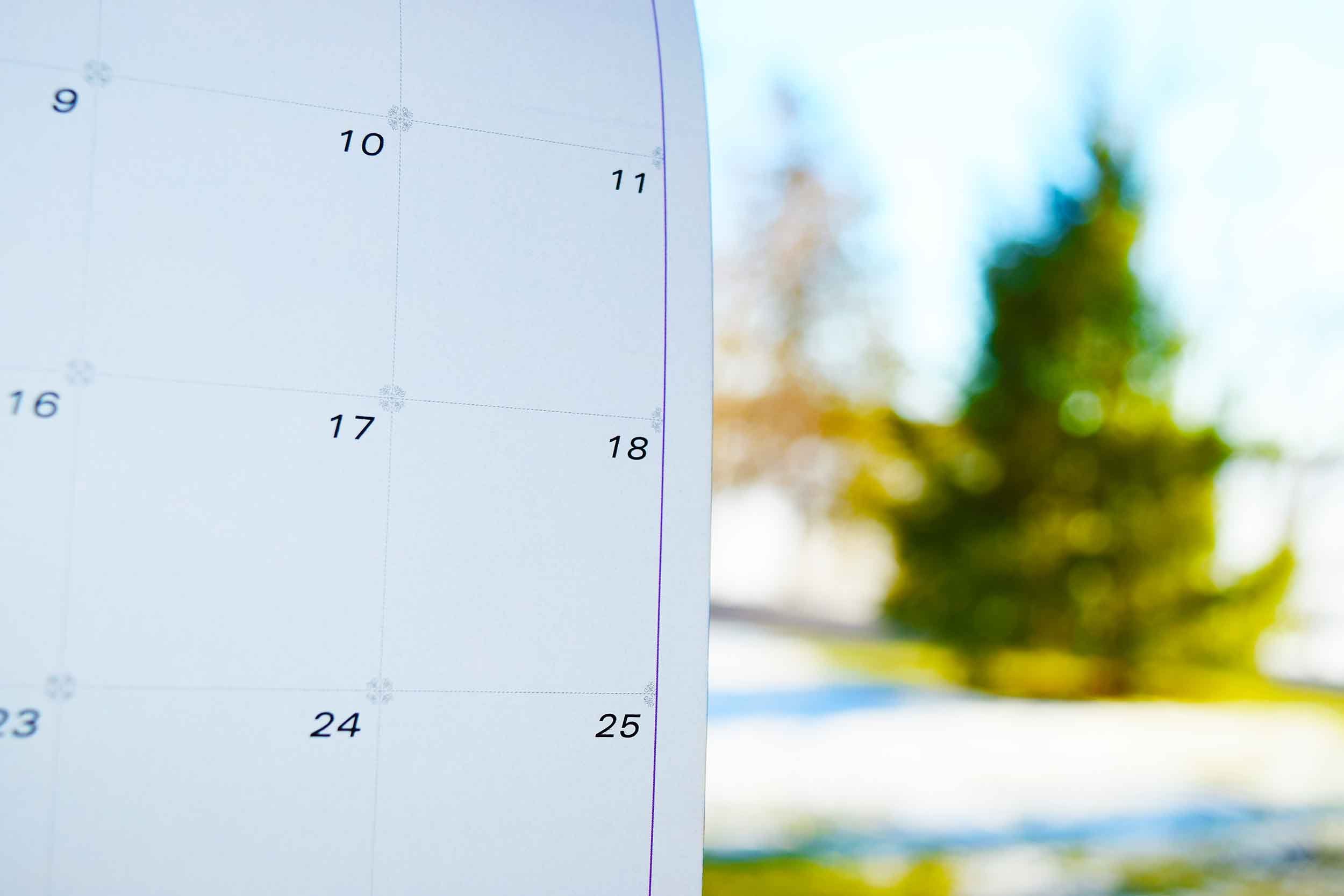Close-up of a paper calendar on the left; to the right is a blurred lawn and evergreen tree with snow visible on the lawn