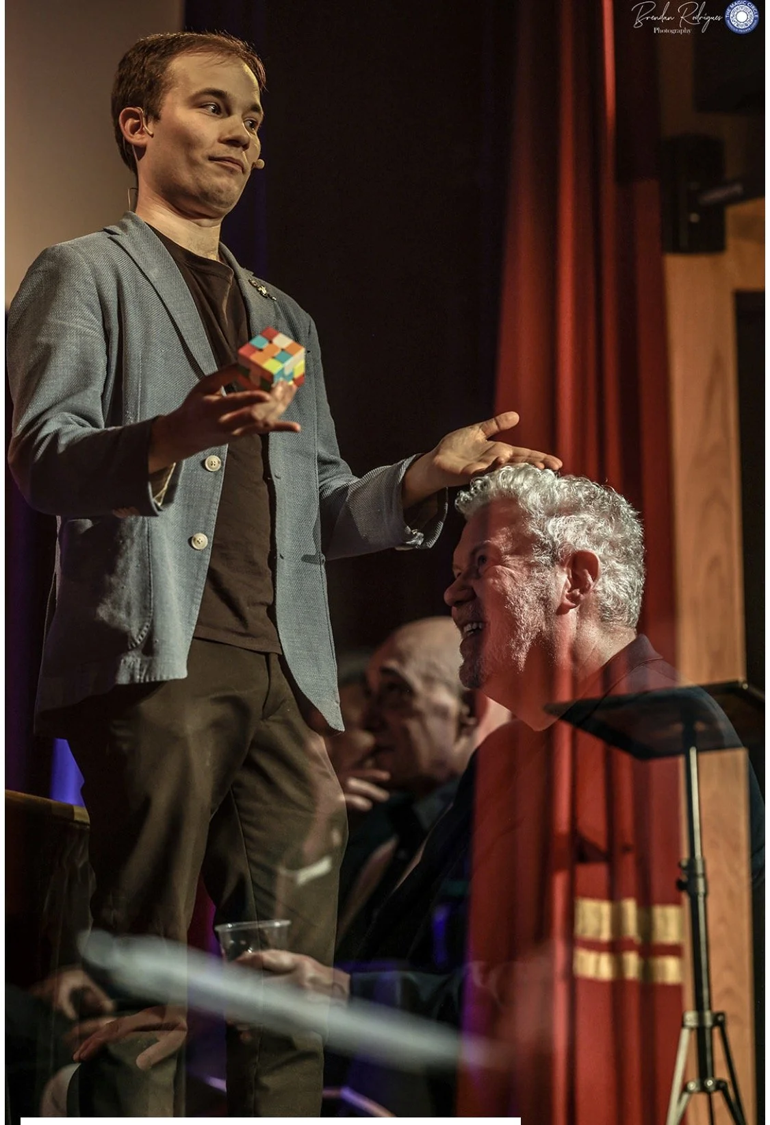 A young man in a gray blazer and black shirt holding a chess piece standing beside an older man with curly gray hair and a beard sitting in a dark blazer, smiling and looking up at the young man. Another man is seen in the background, watching the interaction, with a red curtain and wooden wall behind them.