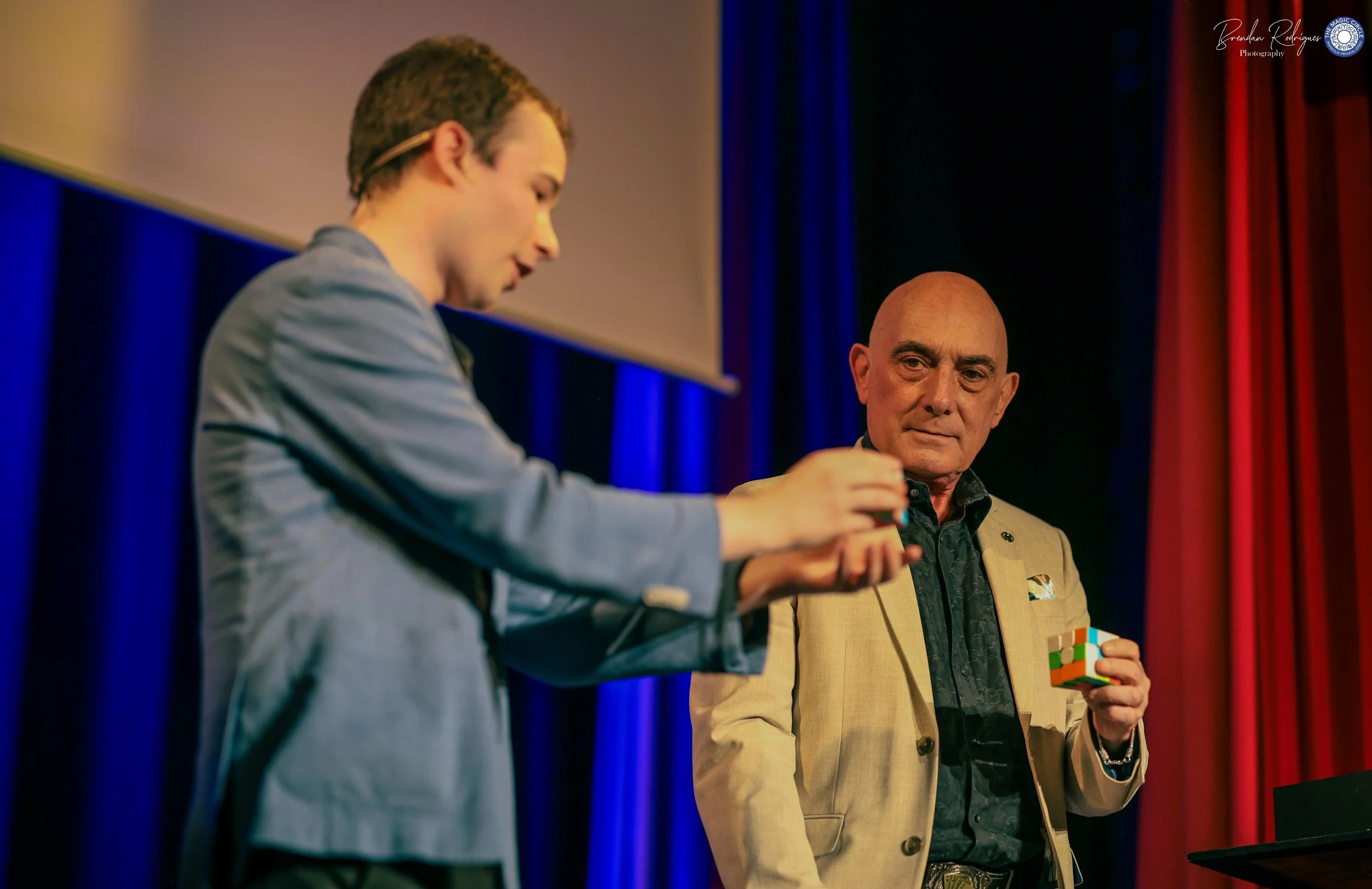 Two men, one with a beard and a mustache, wearing a tan blazer and holding a Rubik's Cube, and the other young man with glasses on his head, wearing a gray blazer, standing on stage at a presentation or event with a blue curtain background.