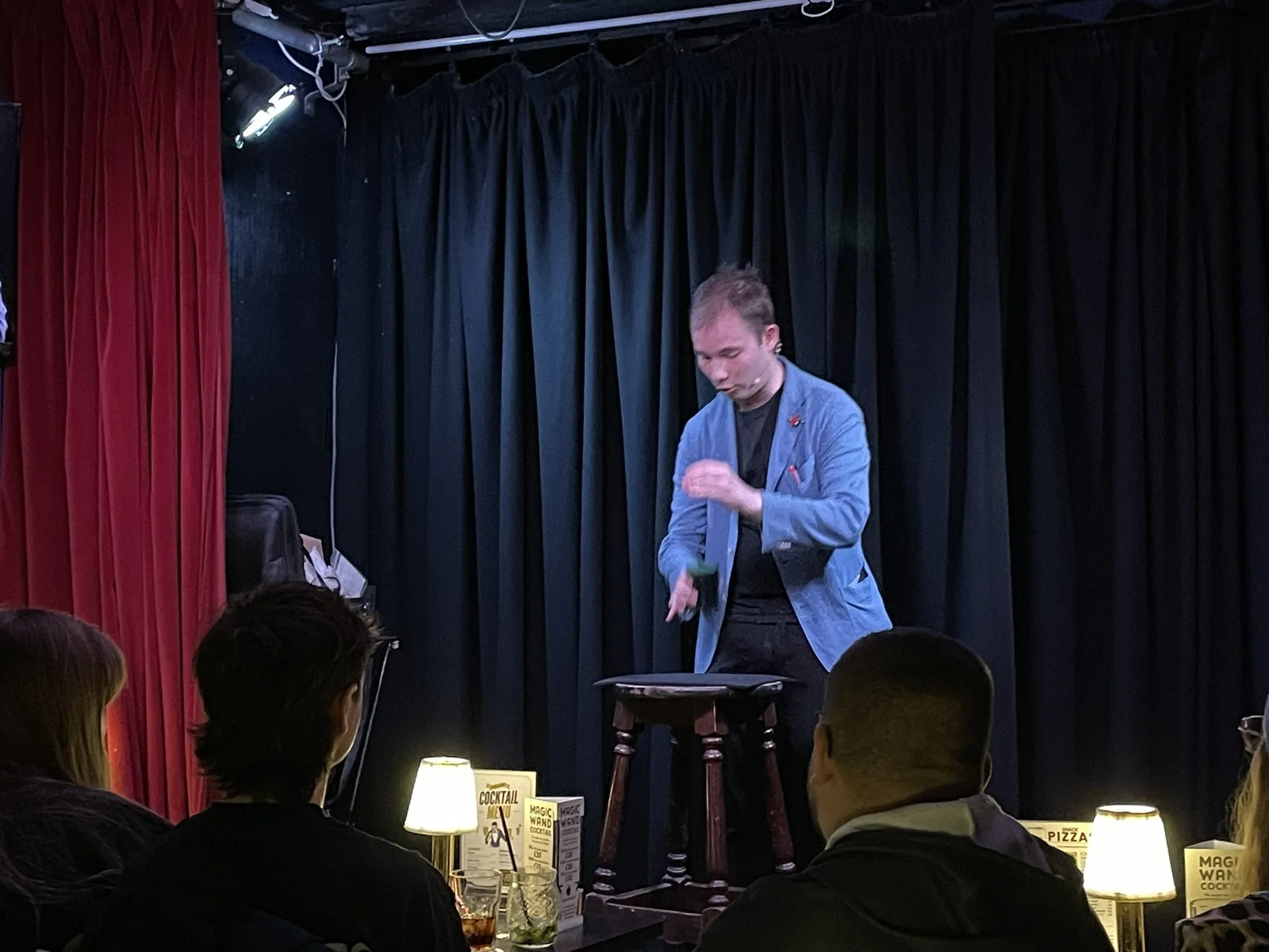 Stand-up comedian performing on stage in a comedy club with black curtains, with audience members watching and lit by small lamps on tables.