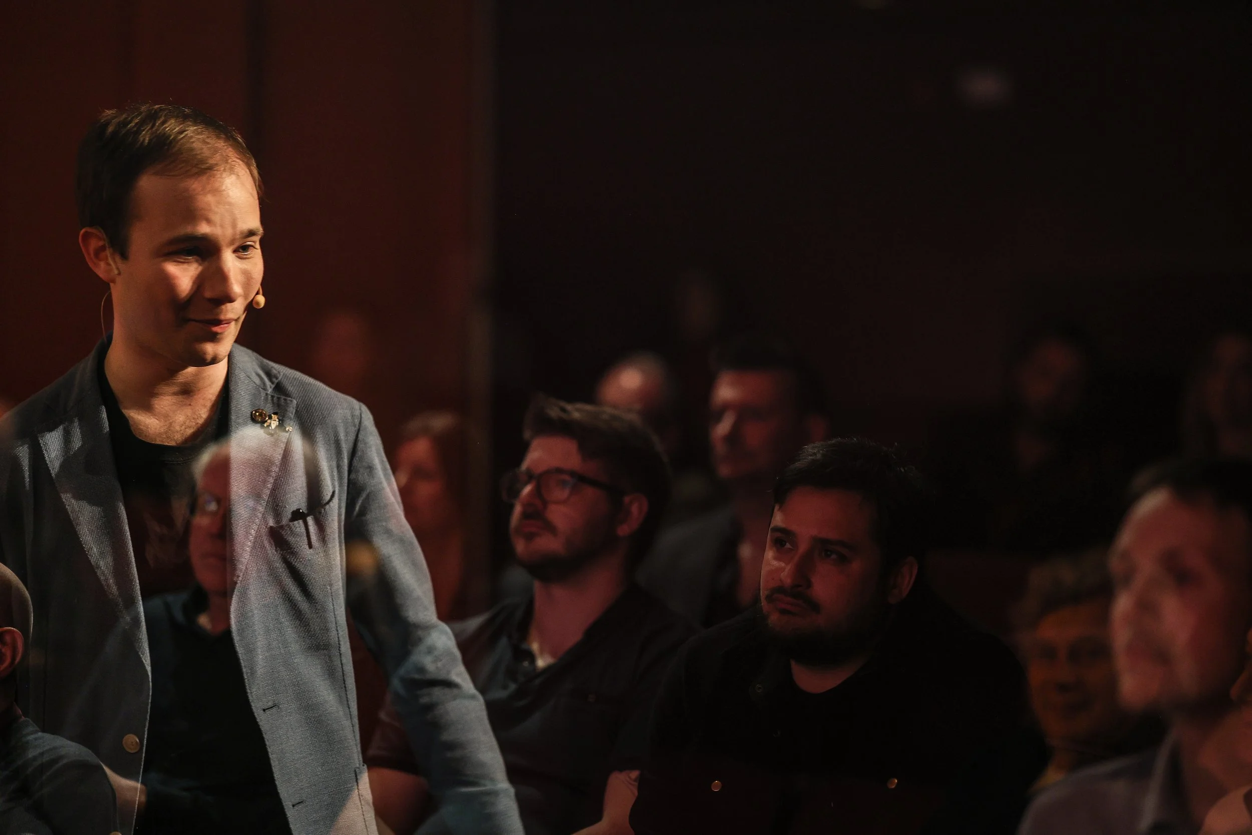 A man in a gray blazer speaking or listening at an event with an audience, some audience members are visible and looking towards him.