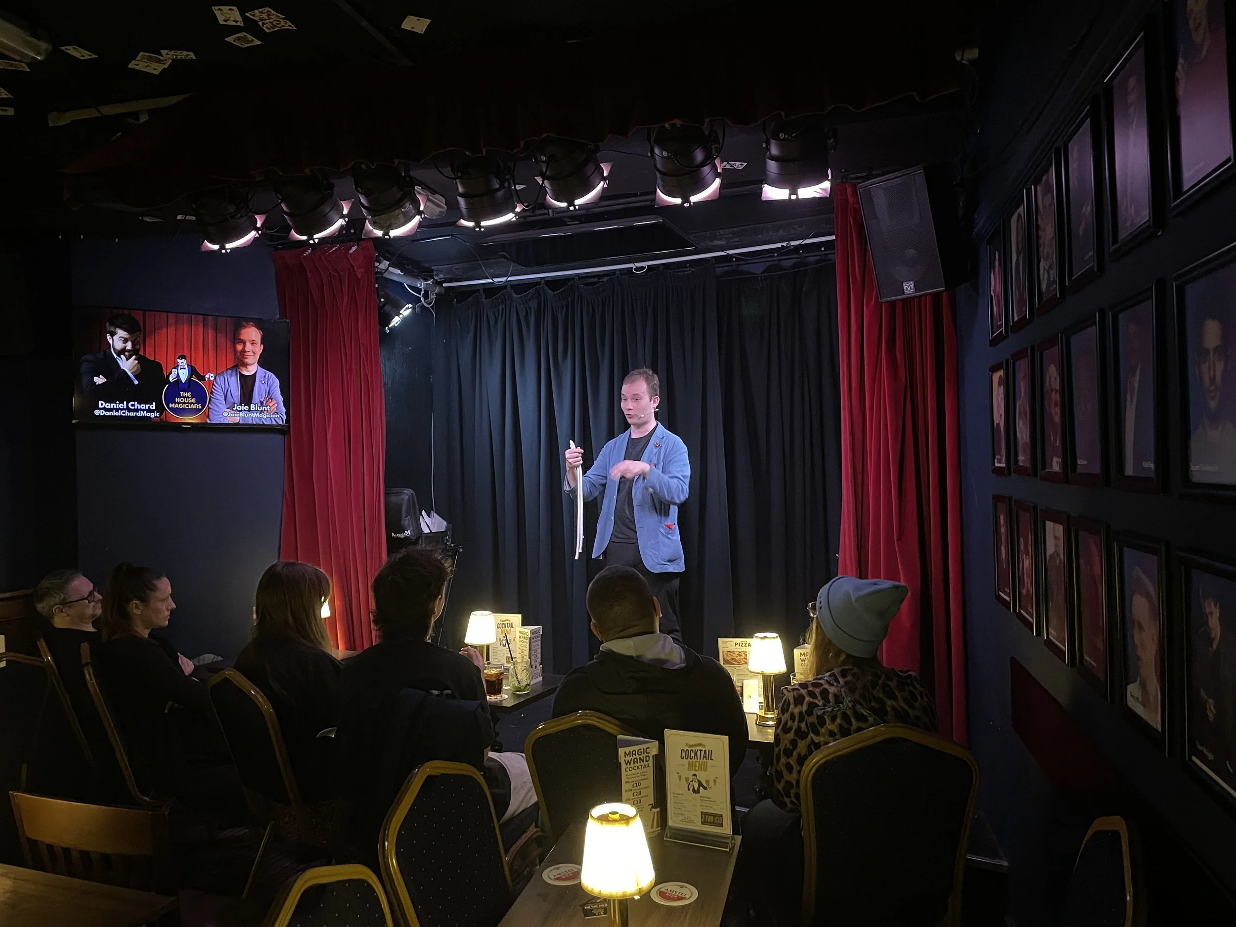 A magician performing on stage in a comedy club, with an audience seated at tables watching. The stage has black curtains and red drapes, with overhead stage lighting. A television monitor on the left shows images of two people, and framed photos are