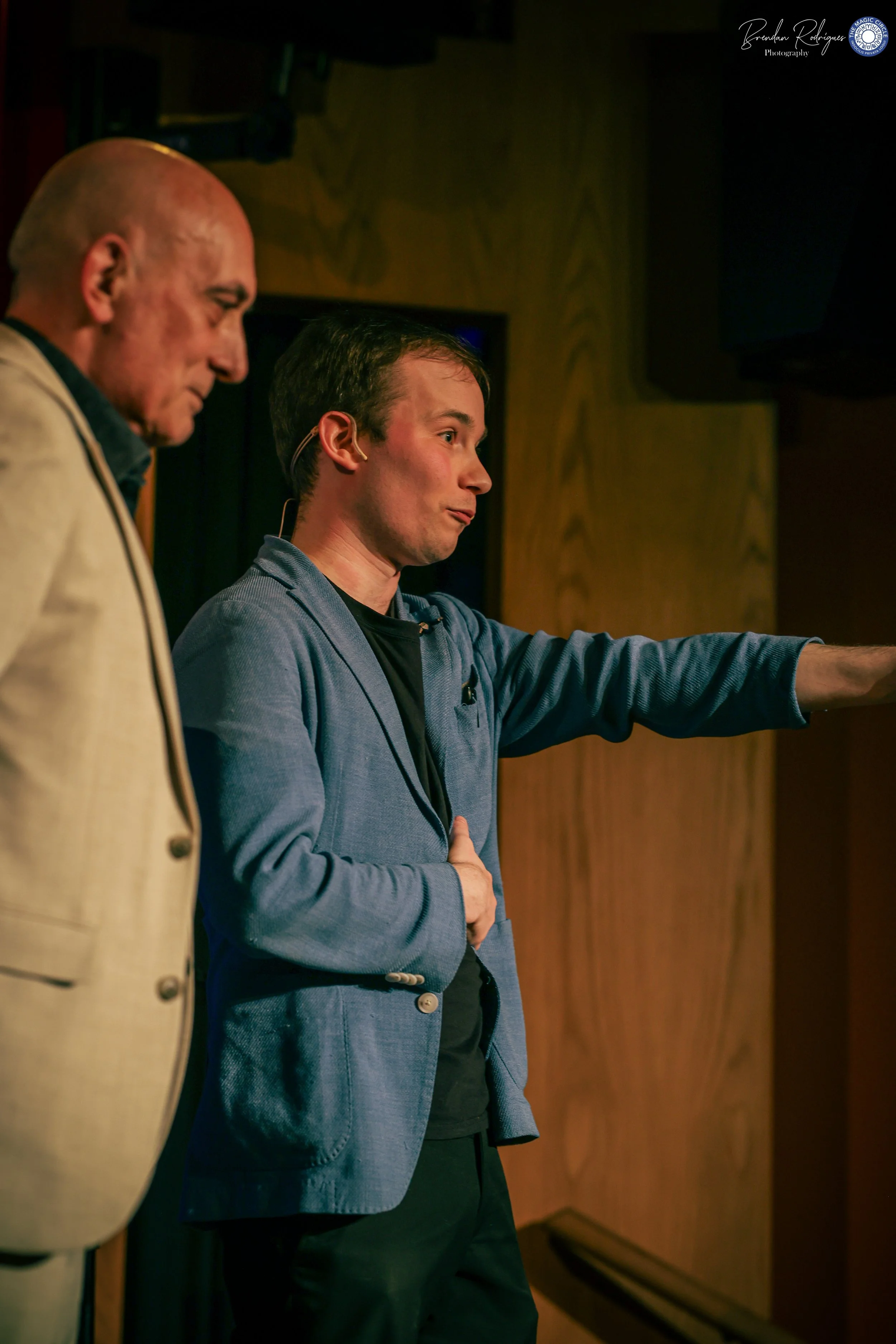 Two men are standing side by side on a stage or platform, with one of them using a microphone headset. The man on the right is making a gesture with his right arm and appears to be speaking or presenting. The background has wooden paneling and electr