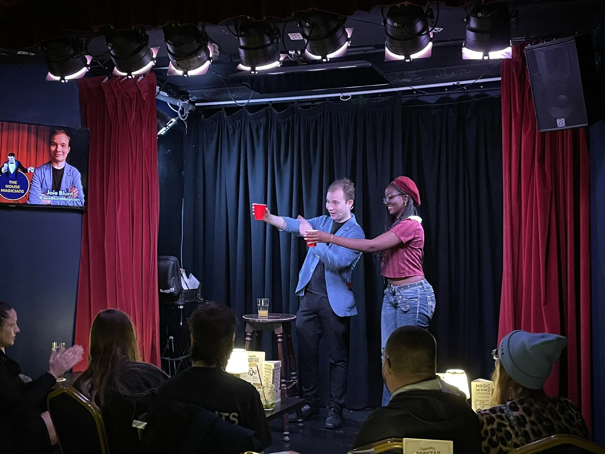 A magician and an assistant performing a magic trick on stage at a comedy club, with an audience watching. The magician is holding a red cup and the assistant is holding a small object, both smiling. The stage has black curtains and red velvet sides,