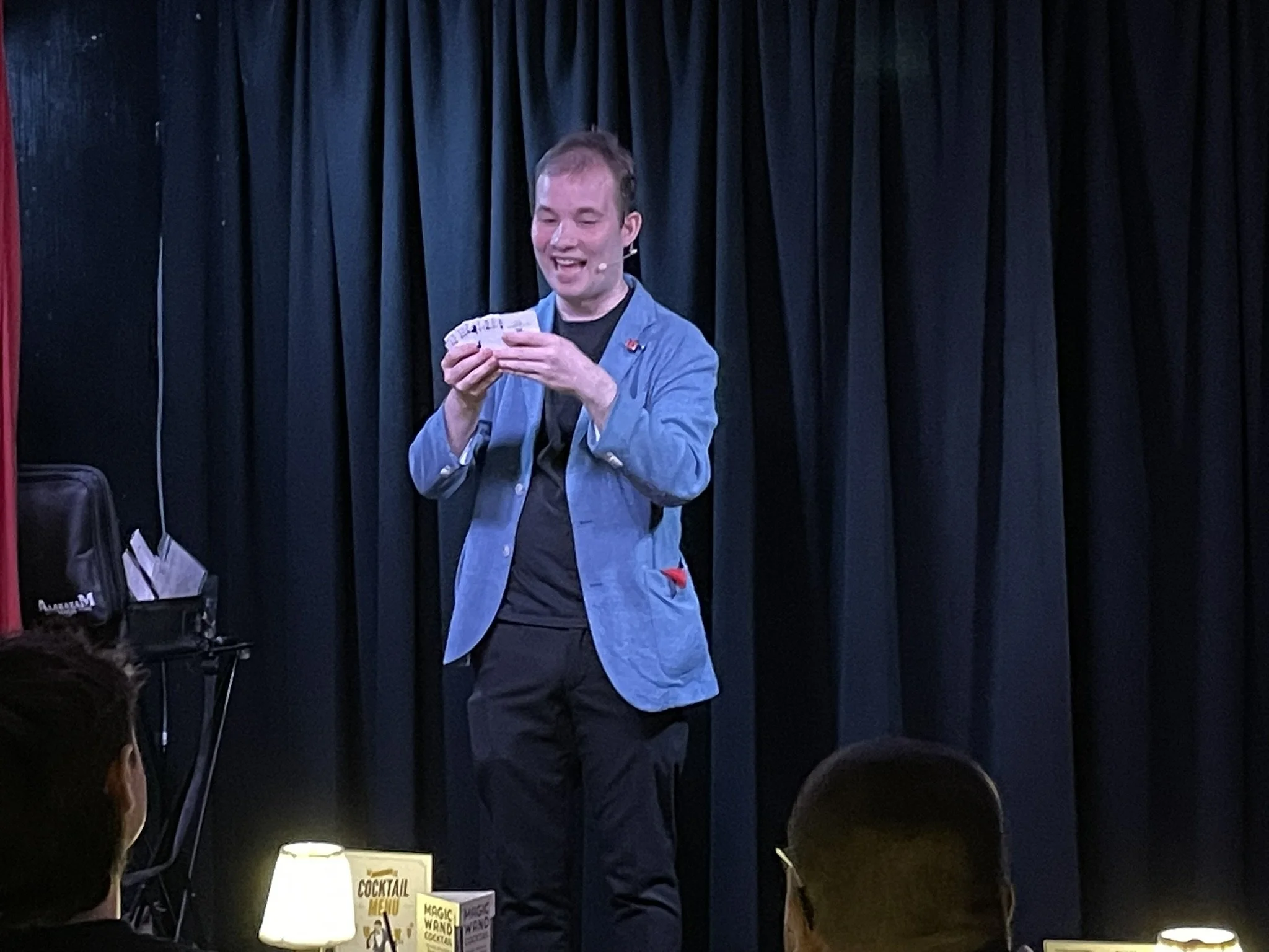 Man performing a magic trick on stage with black curtains, audience visible in foreground.