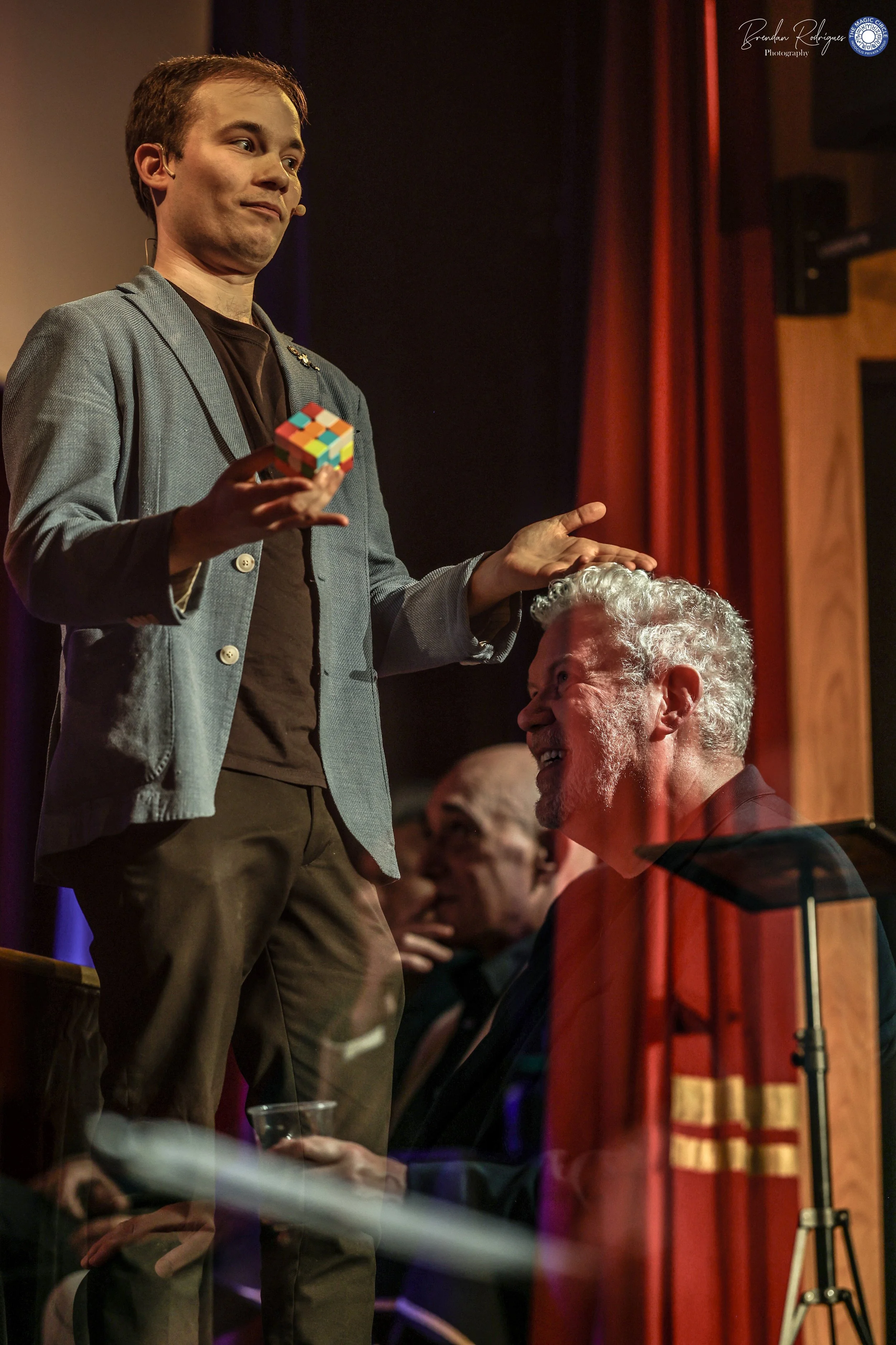 A man with a microphone headset holding a colorful Rubik's Cube on stage, smiling and gesturing with open hands, in front of an audience with a red curtain backdrop.